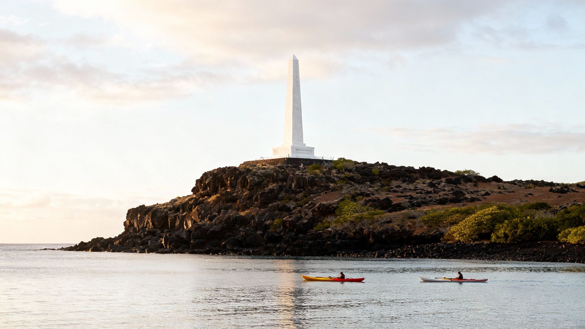 Kayakers paddle near the white Captain Cook Monument on a rocky Hawaiian coast.