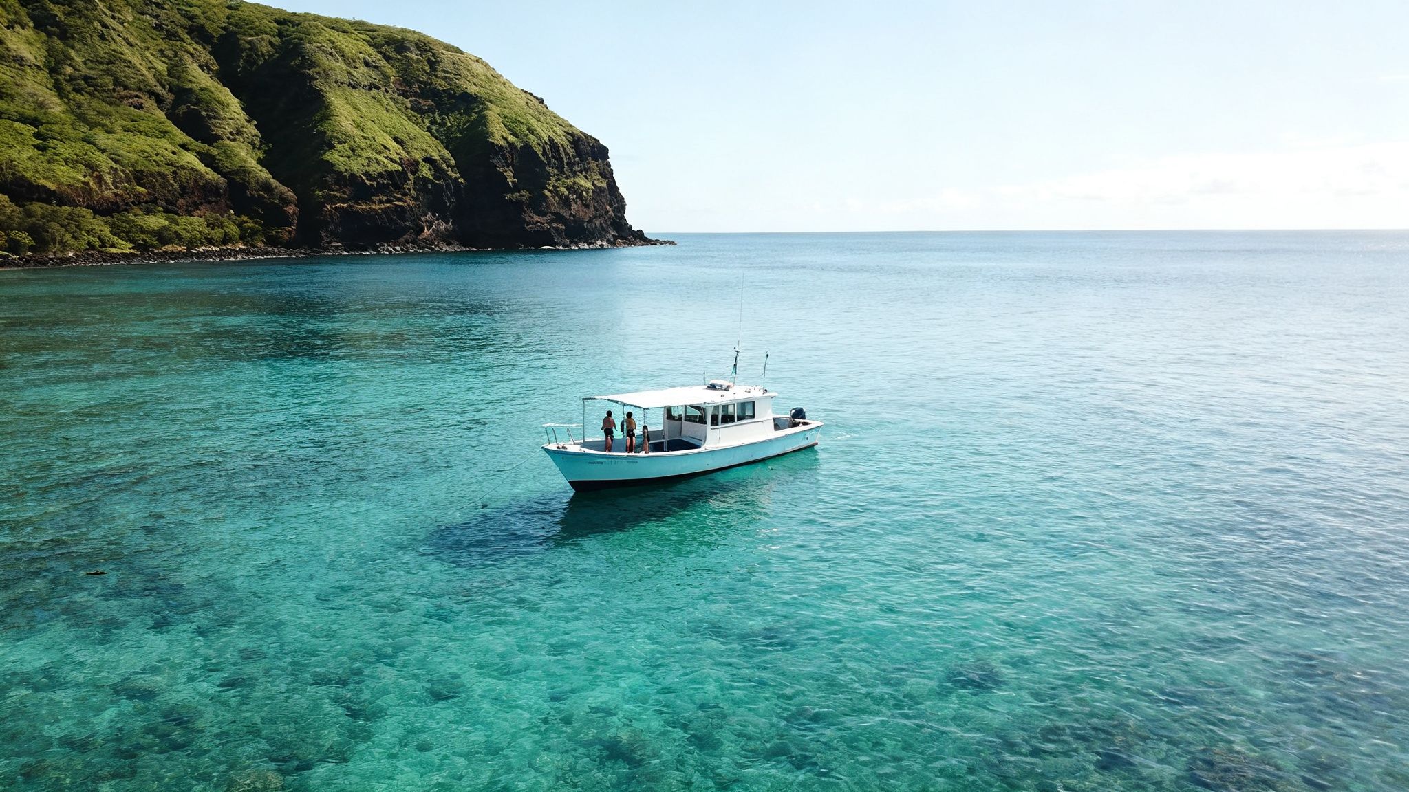 Two people on a boat in clear turquoise water with a green island cliff in Hawaii.