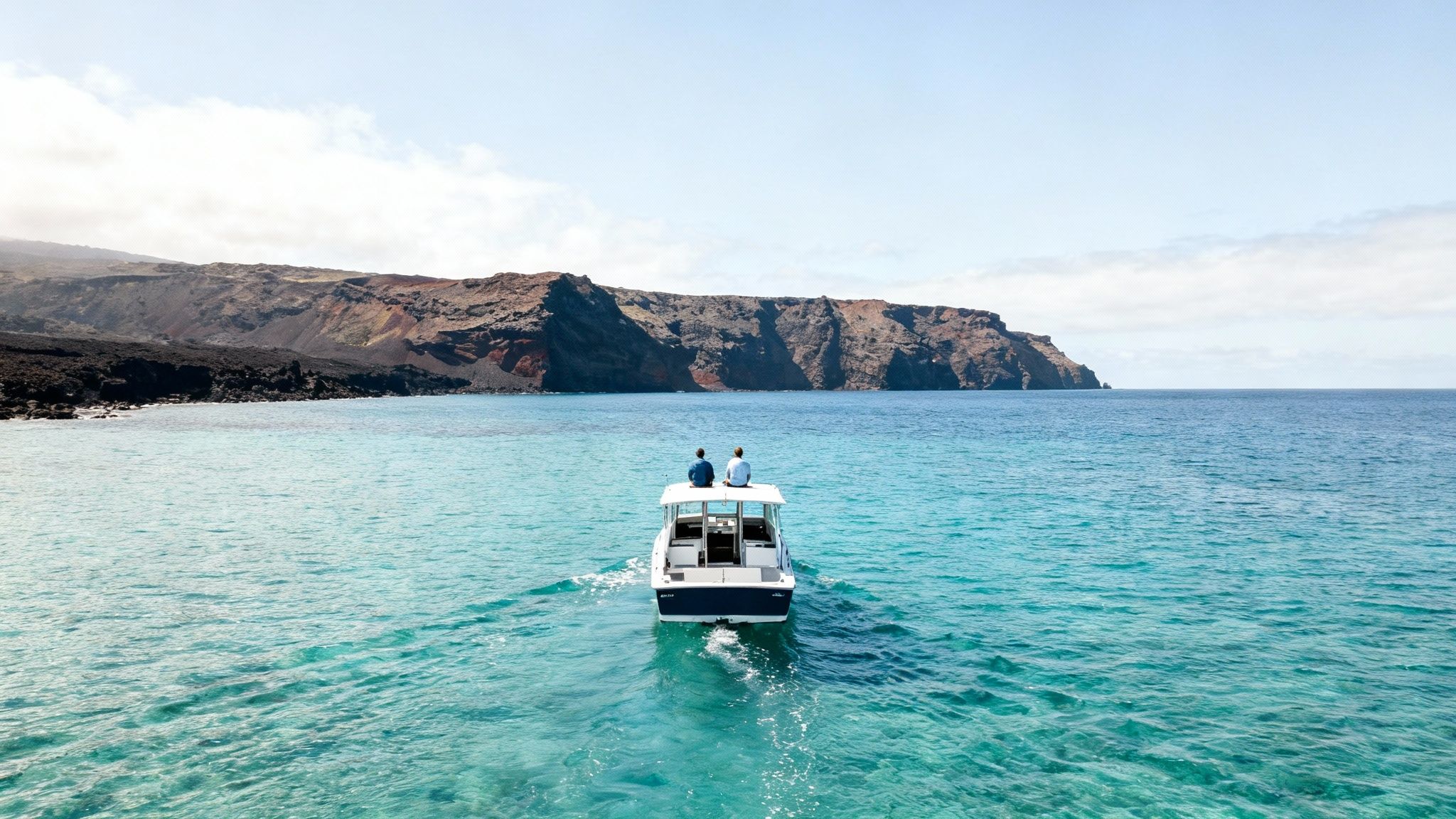 Two people on a small boat cruise on clear blue water towards a rugged, volcanic coastline.