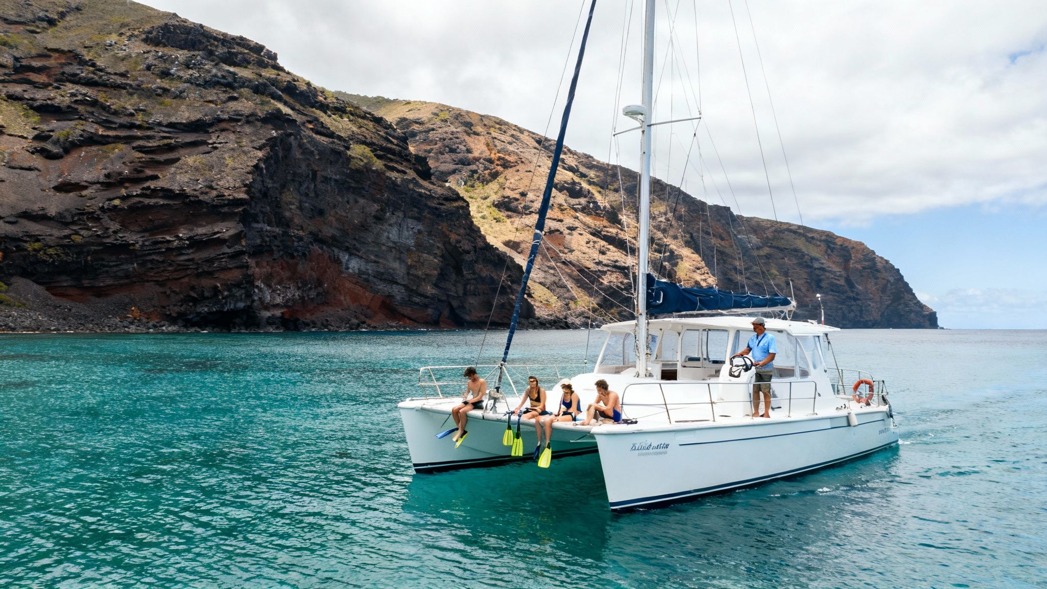 A white catamaran boat carrying people with snorkeling gear on turquoise water near dark rocky cliffs.