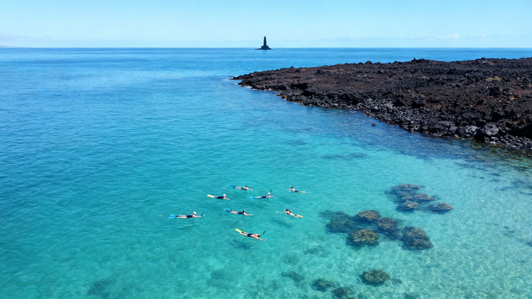 Aerial view of people snorkeling in clear turquoise water near a dark volcanic rock coast with a distant monument.