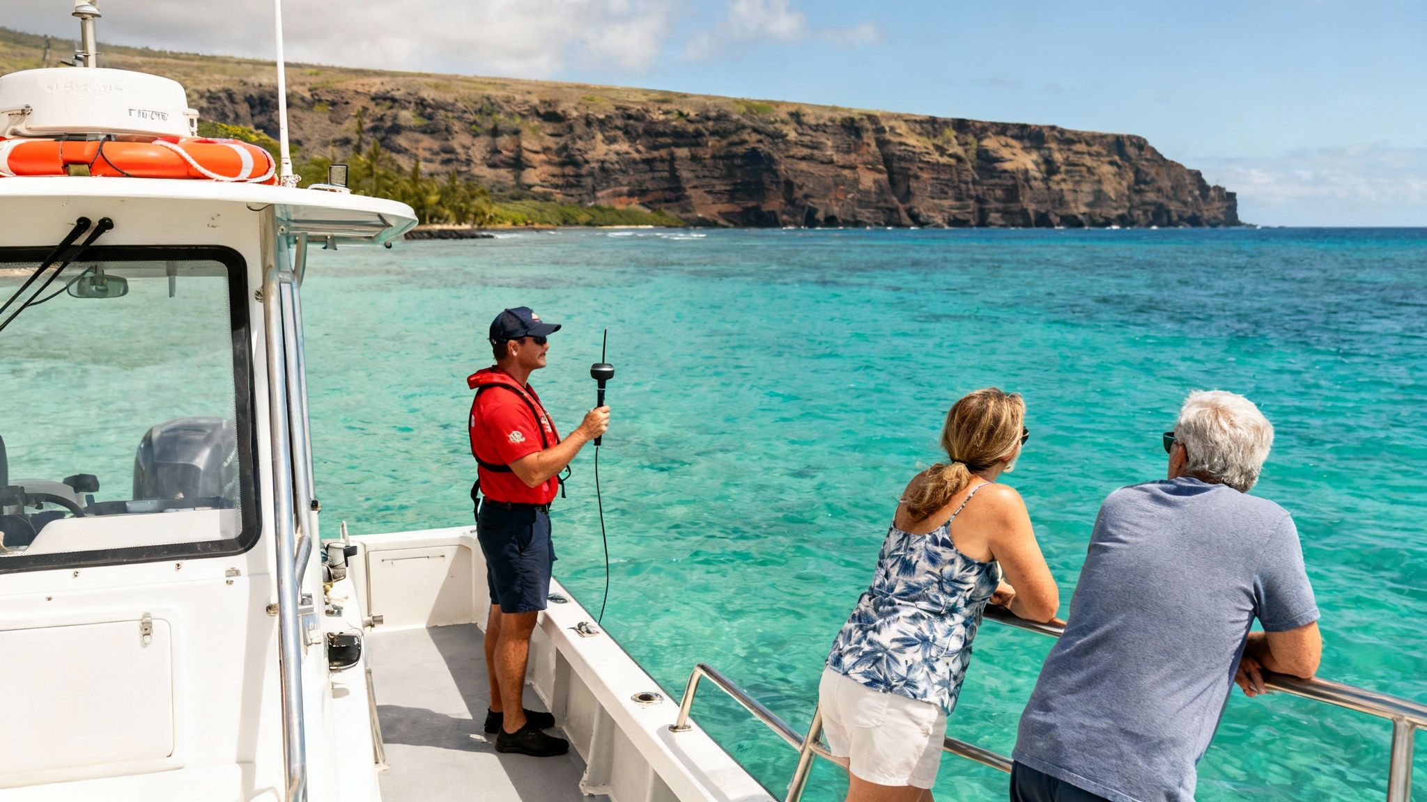 A man on a boat operates equipment while two people admire the clear blue ocean and coastline.