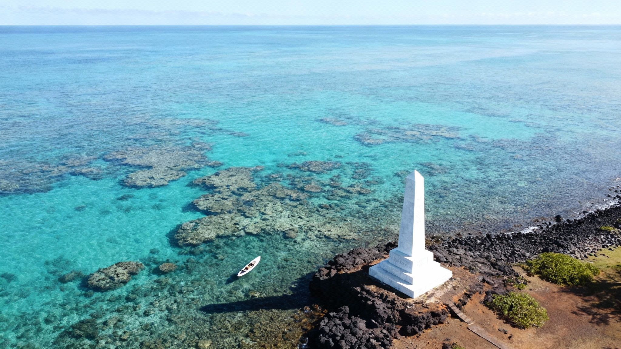 Aerial view of a white obelisk monument on a rocky coast by clear turquoise water with coral reefs and a small boat.