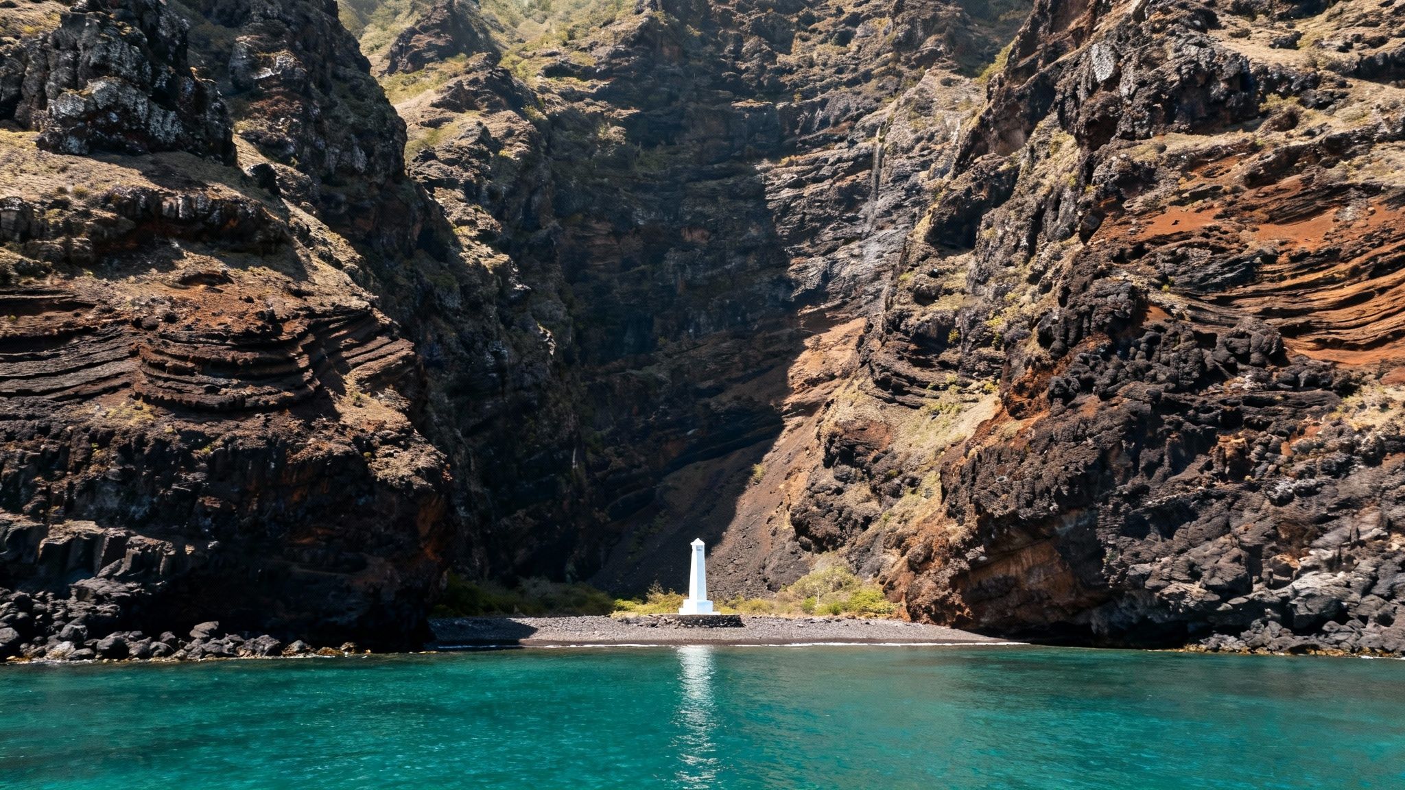 A narrow bay with towering rocky cliffs, turquoise water, and a white monument on a pebble beach.