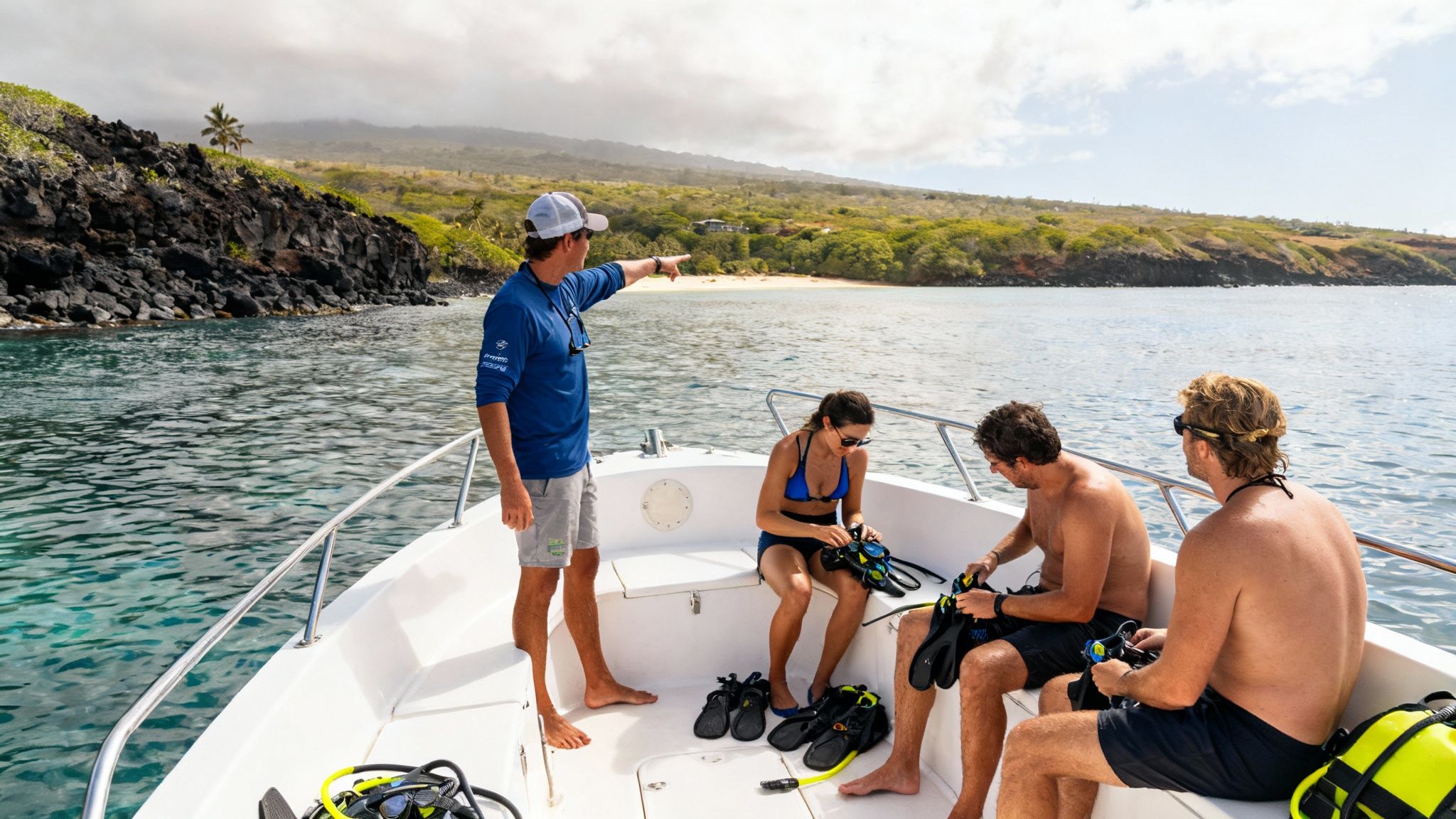 A boat captain points towards a sandy beach as tourists prepare snorkel gear.