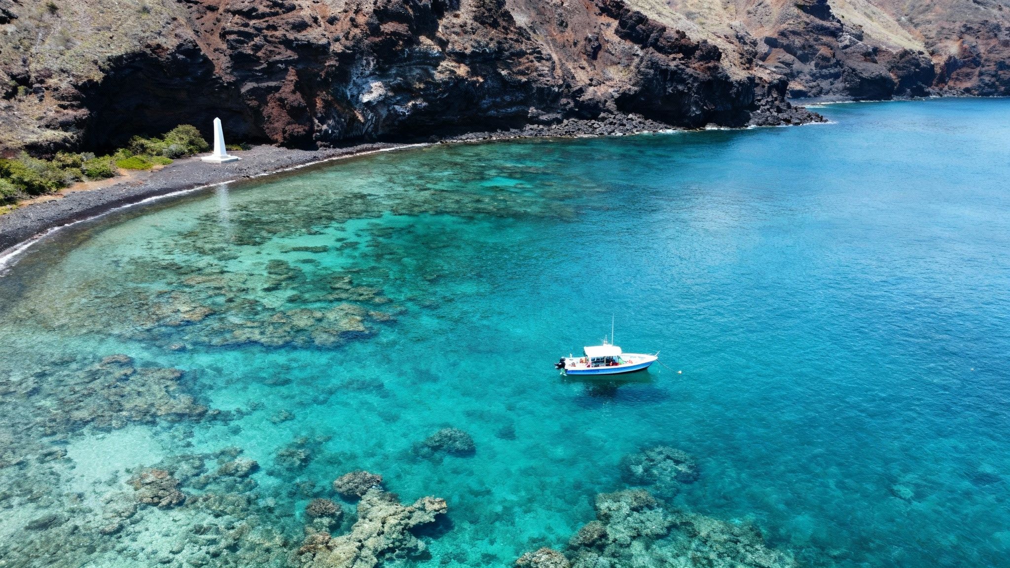 Aerial view of a white monument on a black pebble beach with a boat in clear blue water and coral.