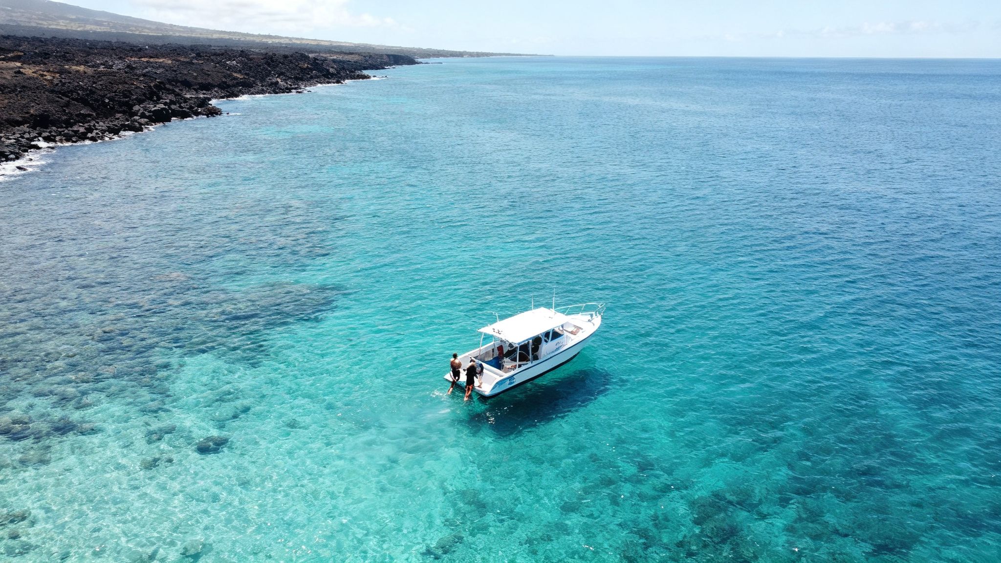 Aerial view of a a white boat with people in clear turquoise water near a rocky coastline.