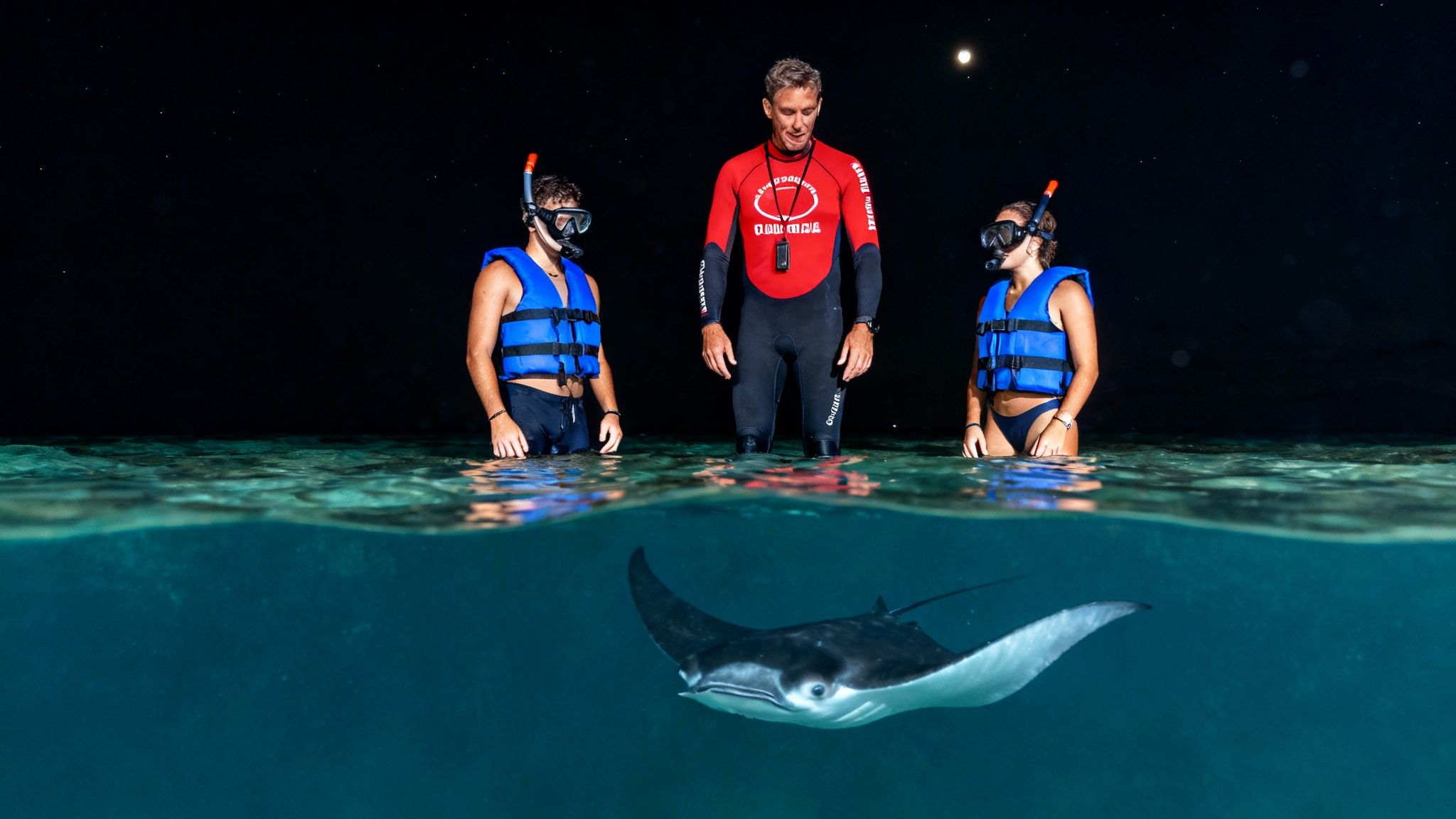 A guide and two snorkelers observe a graceful manta ray in clear water at night.