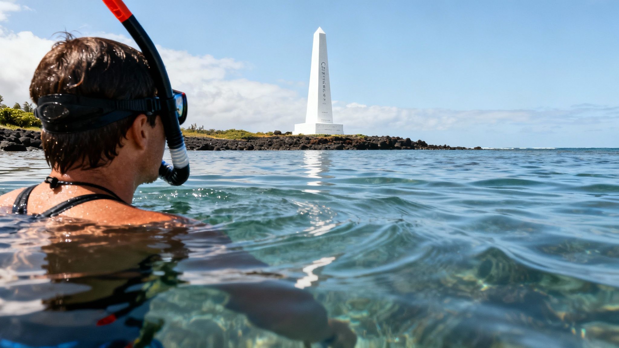 A person in snorkeling gear looks from clear ocean water towards a tall white monument on a rocky shore.