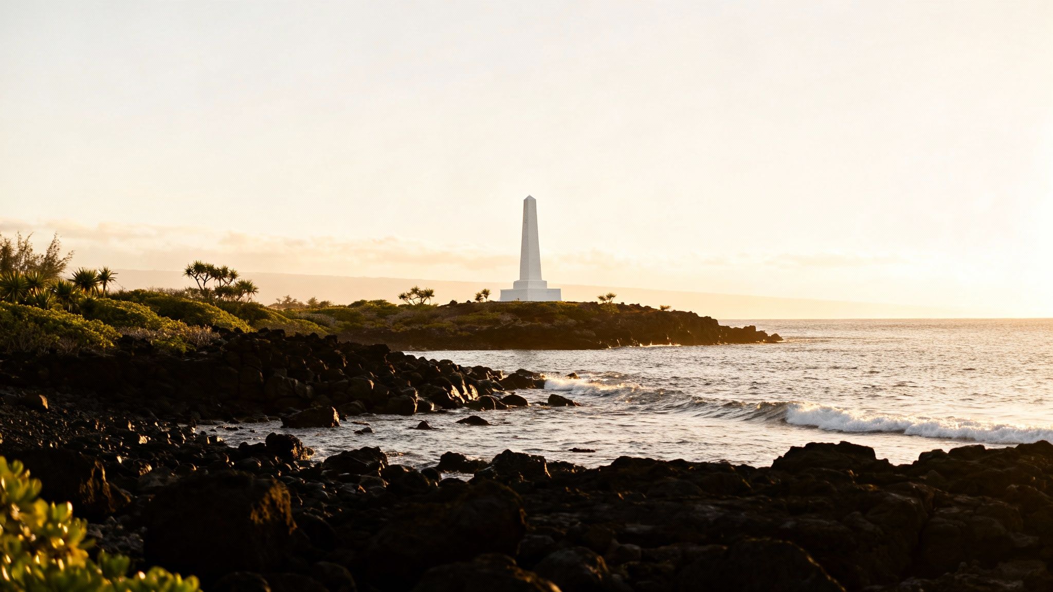 A white obelisk monument stands on a rocky coastline with green foliage, ocean waves, and a golden sunset sky.