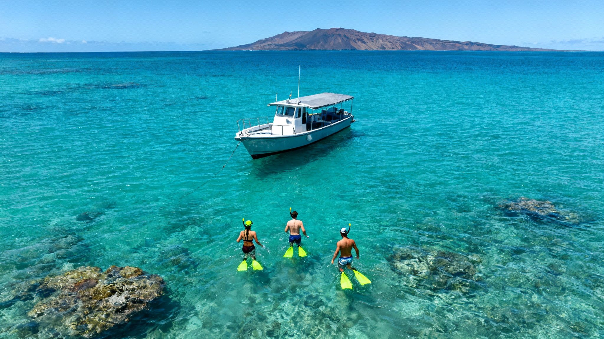 Three snorkelers with fins walk in clear turquoise water towards a boat, with a distant tropical island.