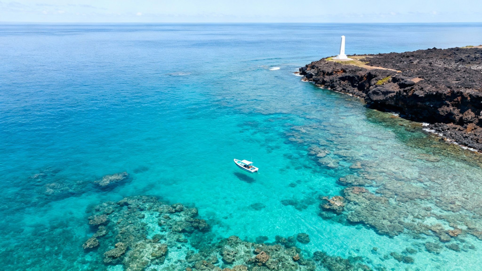 Aerial view of a boat in clear turquoise water near a volcanic coastline with a white lighthouse.