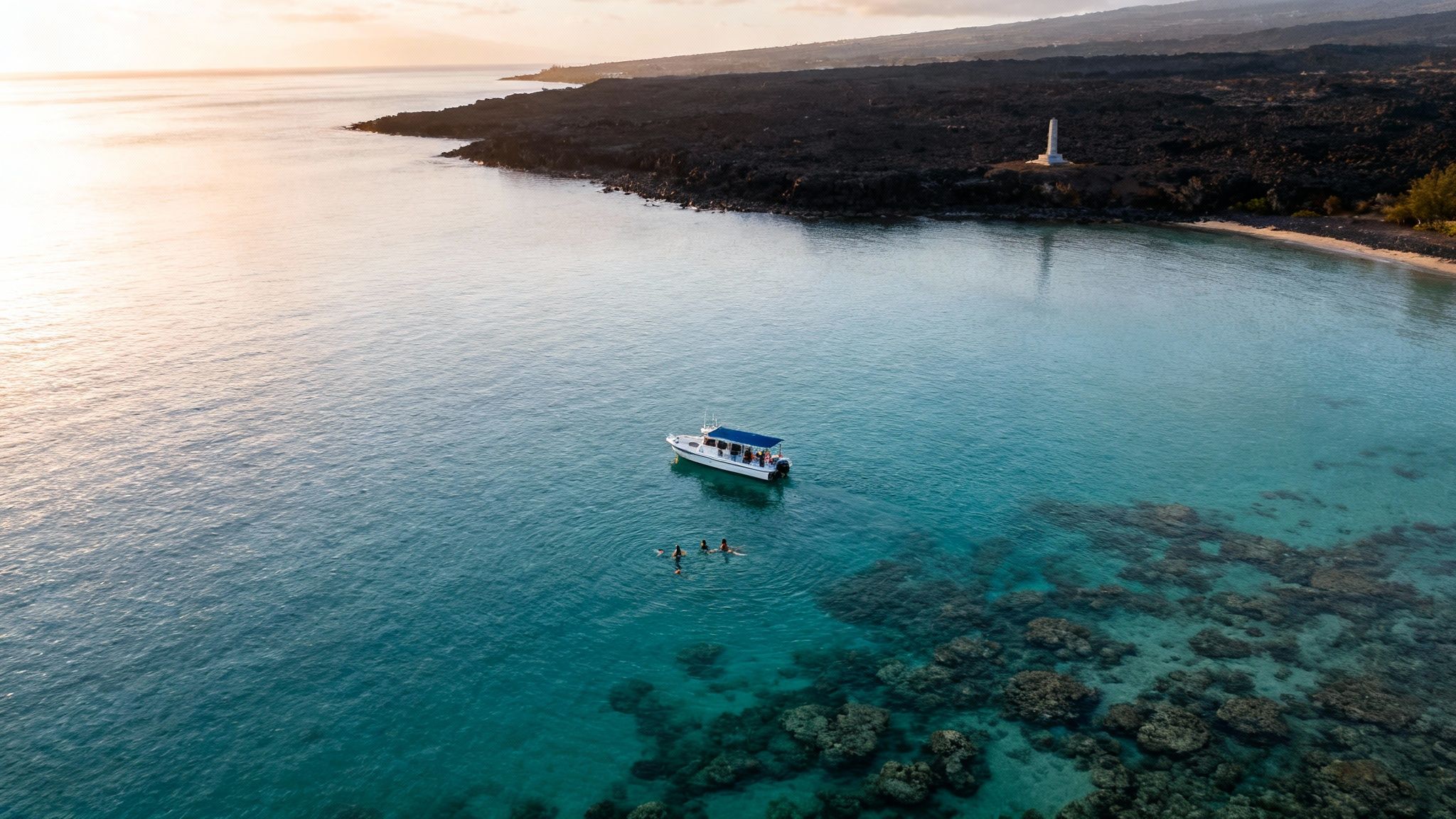 Aerial view of a boat and snorkelers near a tropical coastline with a white monument and lava rocks.