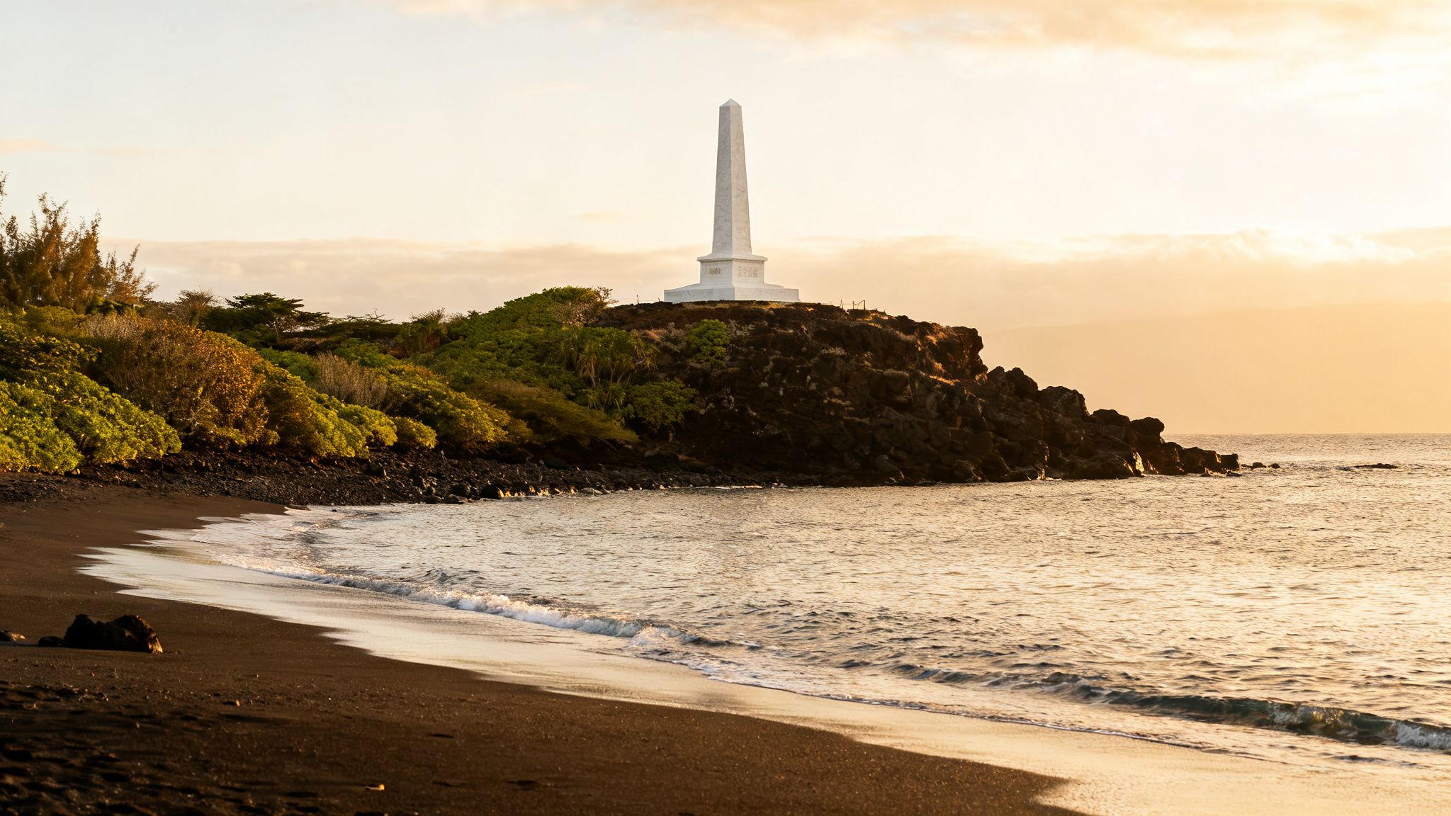 A white obelisk monument stands on a rocky cliff overlooking a black sand beach and ocean at sunset.
