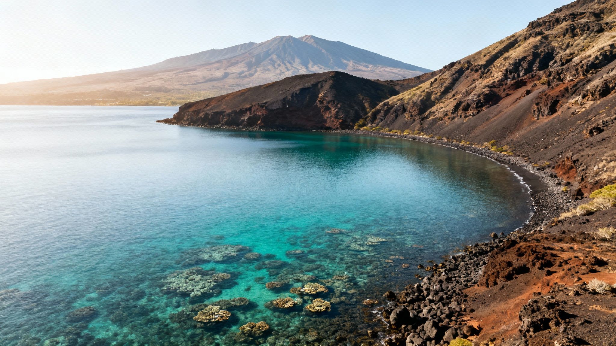 Aerial view of a volcanic coastline with clear turquoise water, coral reefs, black sand beach, and mountains.