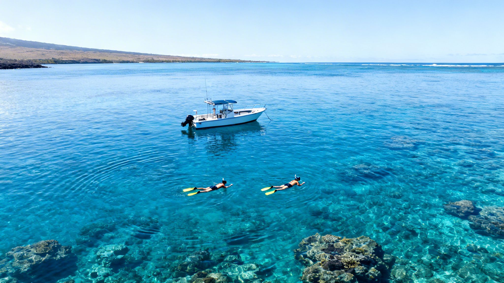Aerial view of two people snorkeling in clear blue Hawaiian waters near a boat and coastline.