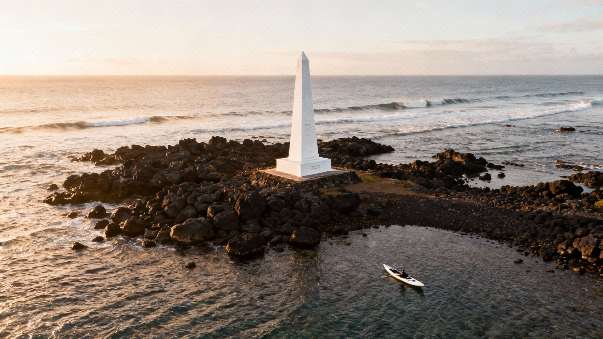 A striking white monument stands on the shore of a clear blue bay, surrounded by lush green cliffs.