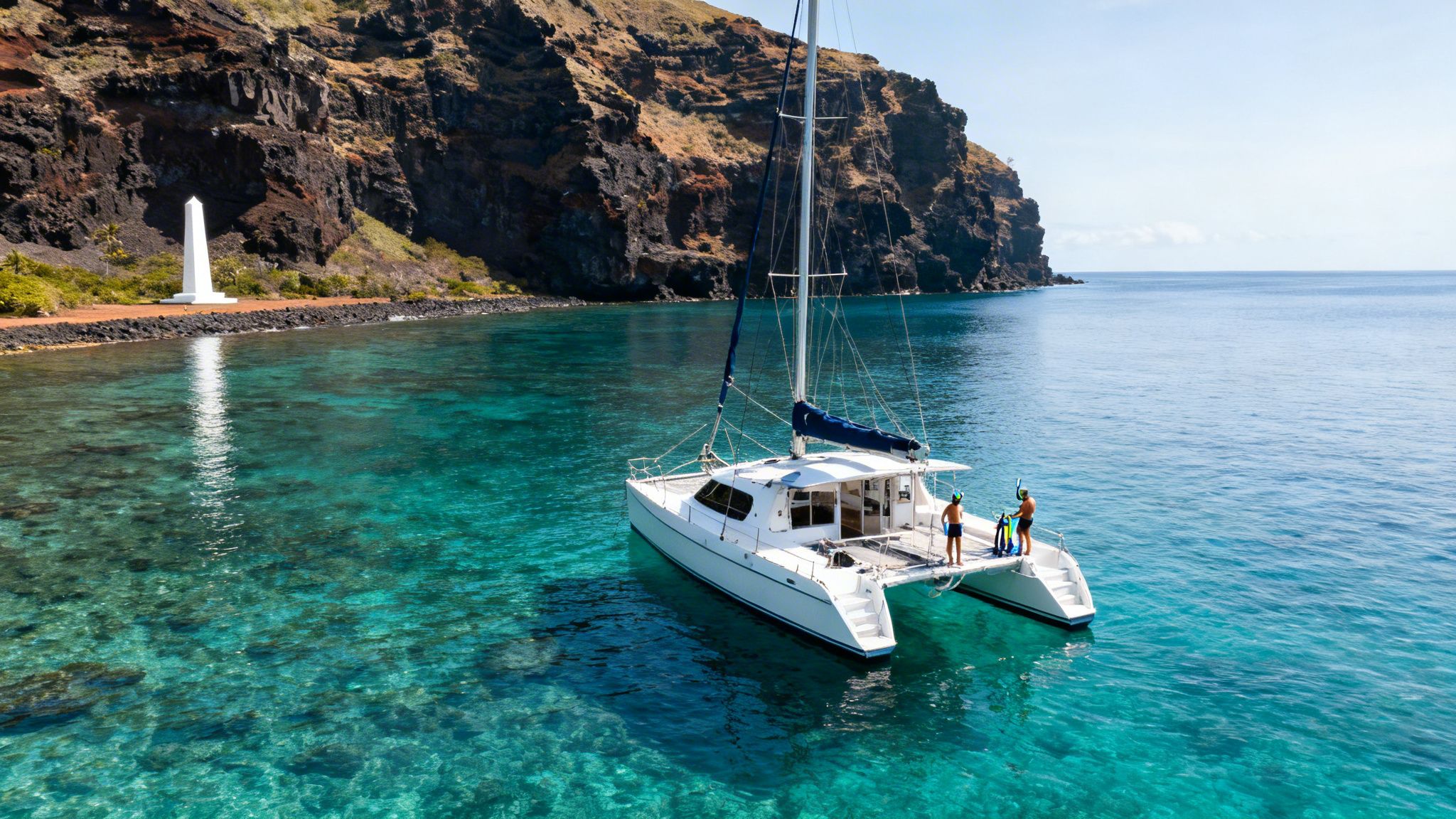 A catamaran is anchored in clear turquoise water near a steep cliff, with two men and an obelisk.
