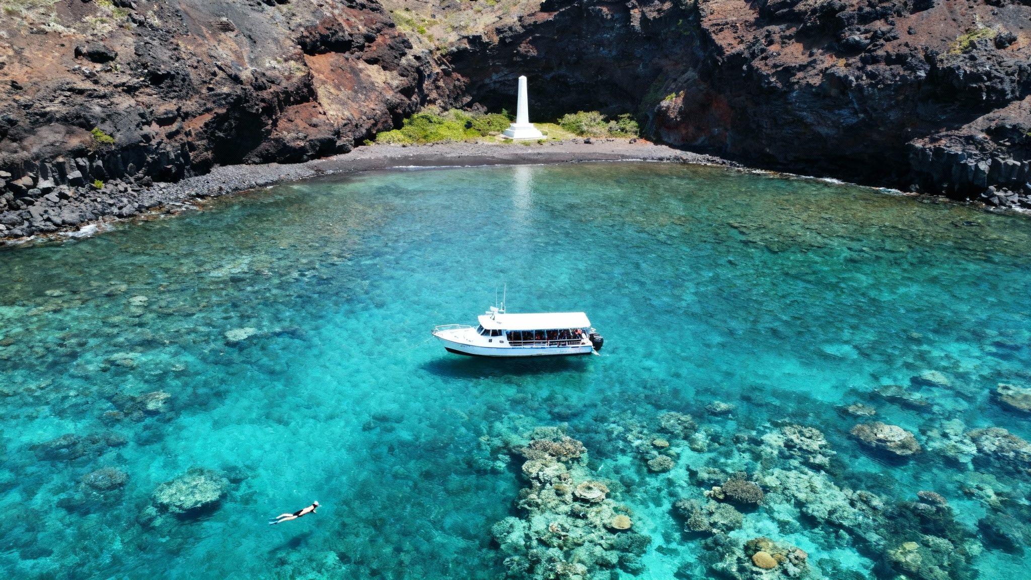 Aerial view of a boat in turquoise water, a snorkeler, a rocky coast, and a white monument.