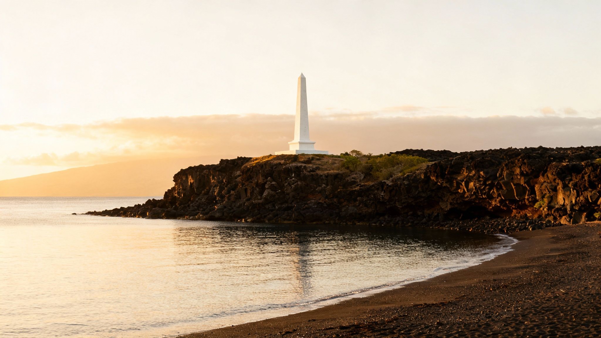 A tall white obelisk monument stands on a dark volcanic cliff overlooking a calm bay at sunset.