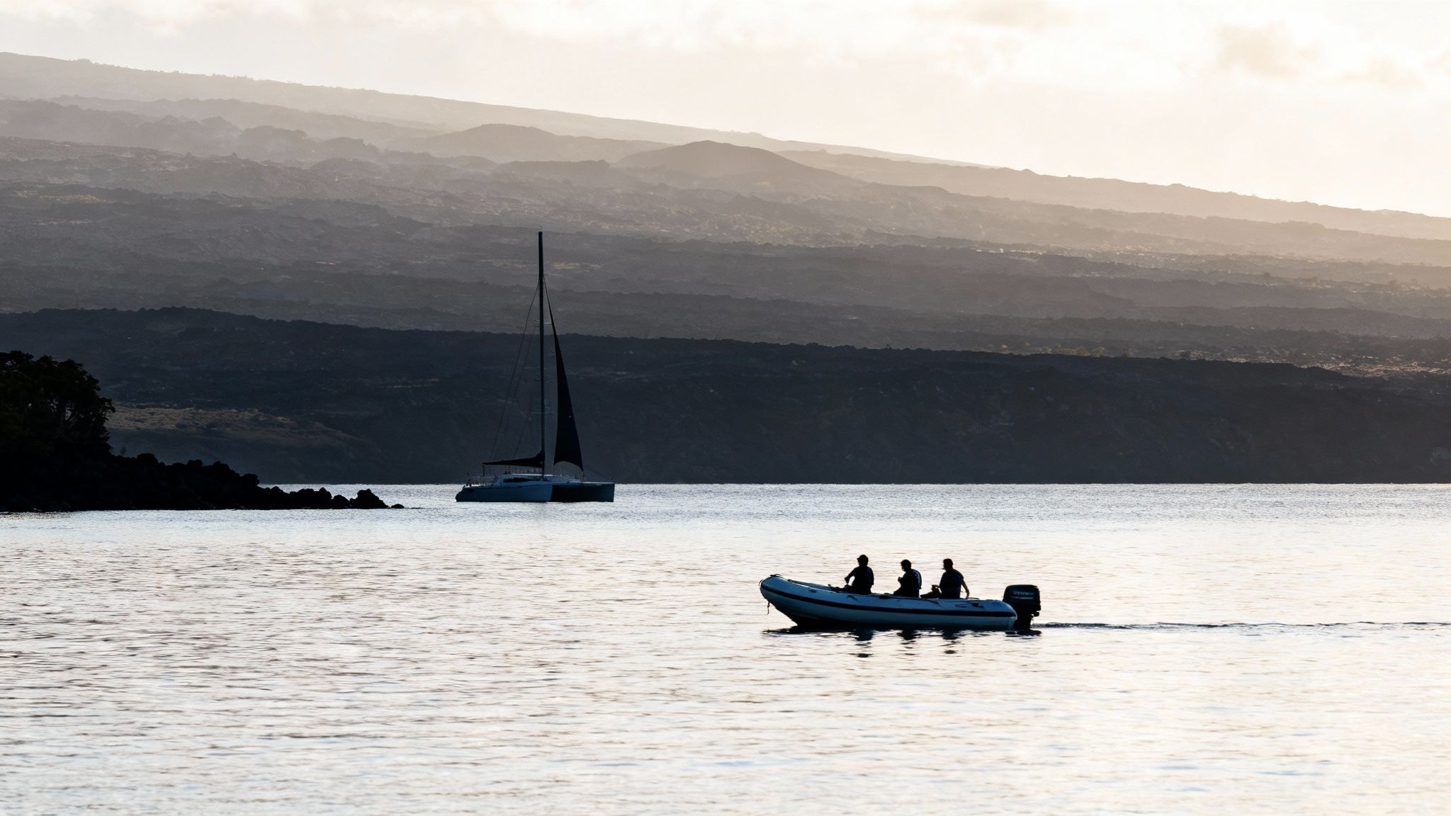 Silhouetted boats on calm water with volcanic mountains in the background at sunset.