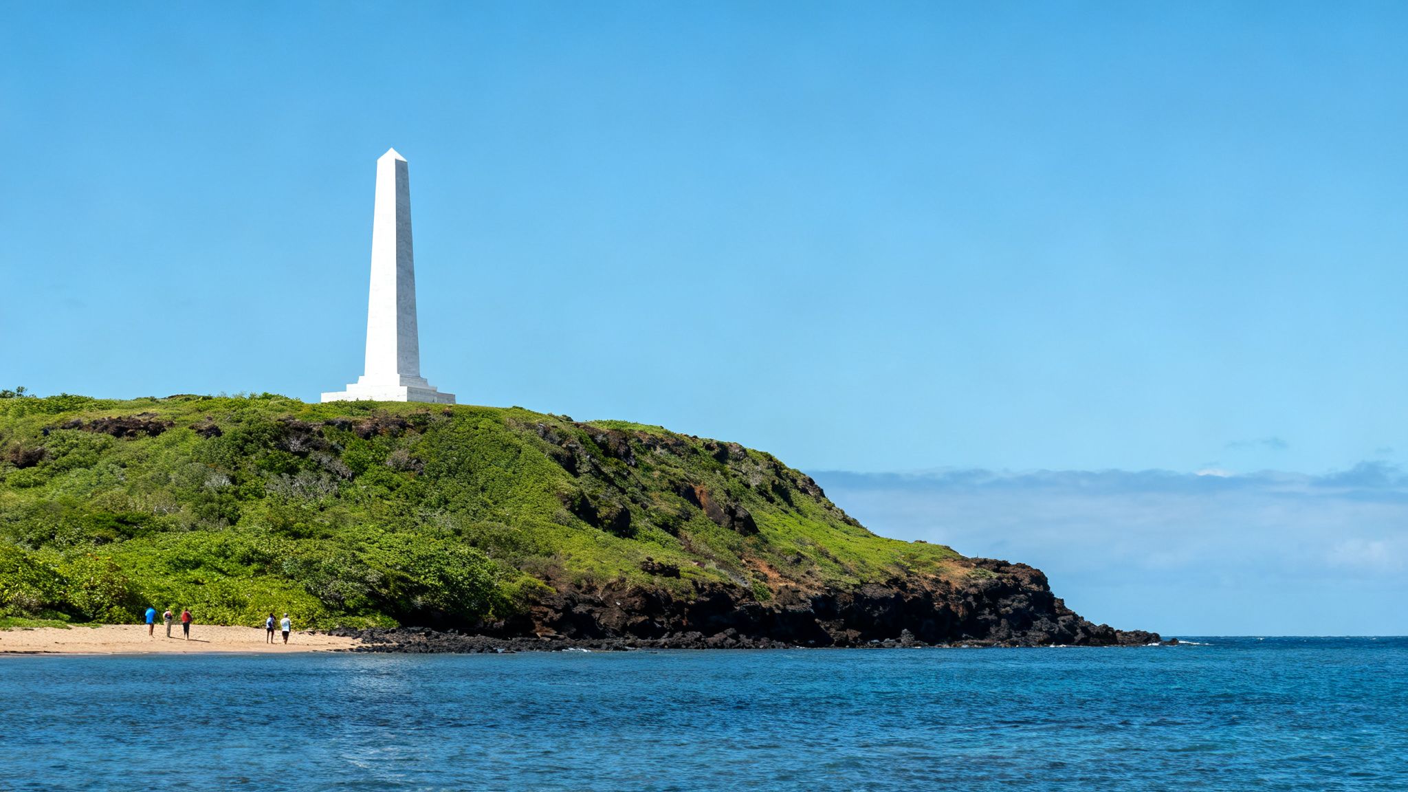 A white obelisk monument stands tall on a green hill overlooking a blue ocean and sandy beach.