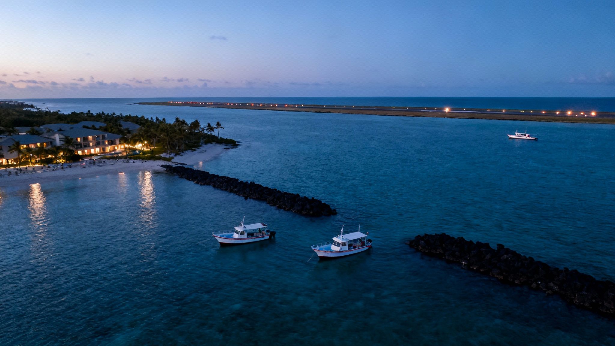 Aerial view of a coastal resort at dusk with boats in the ocean and an illuminated airport runway in the distance.