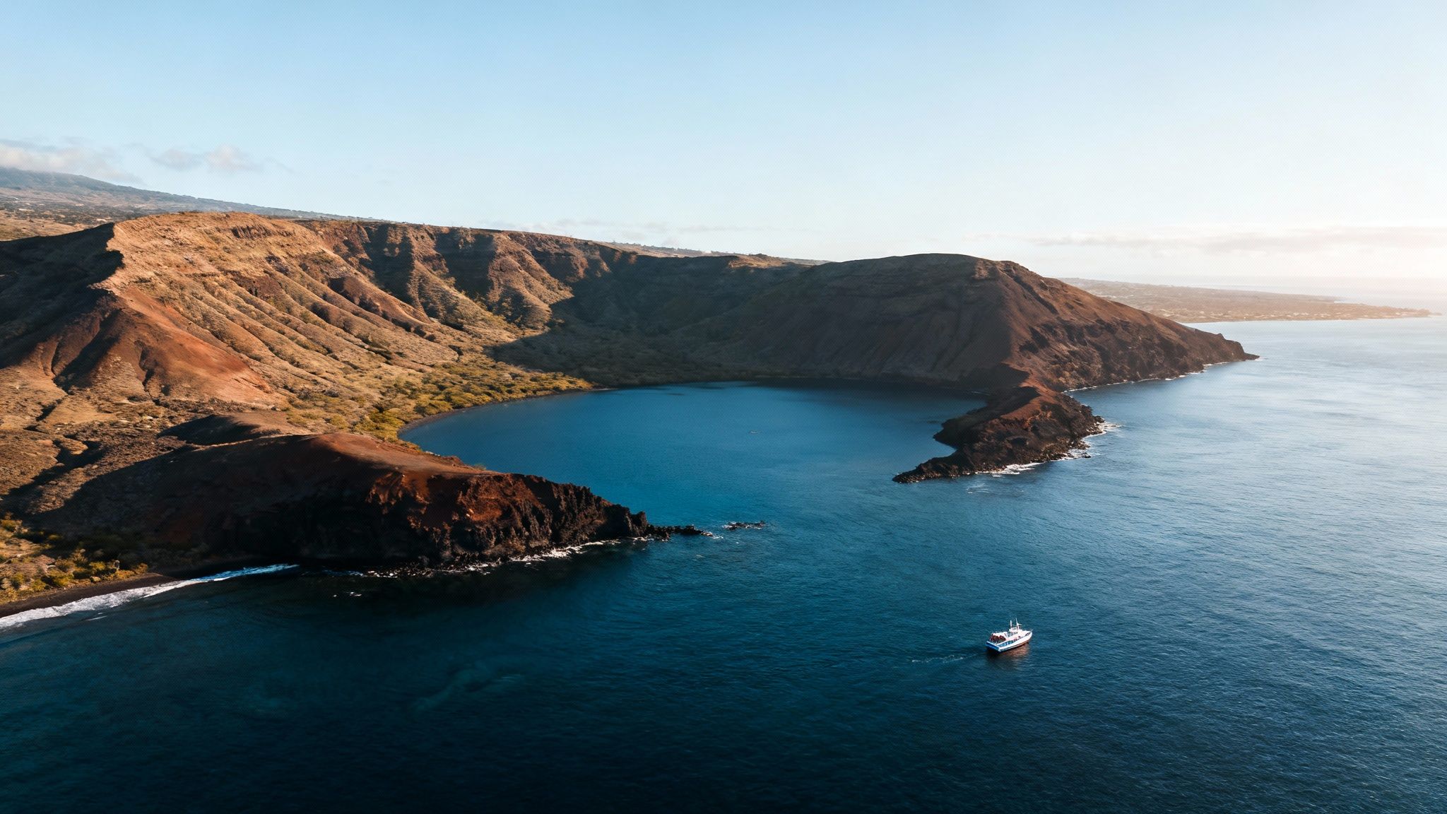 A white boat cruises in a dark blue volcanic bay with rugged, sunlit coastal cliffs.