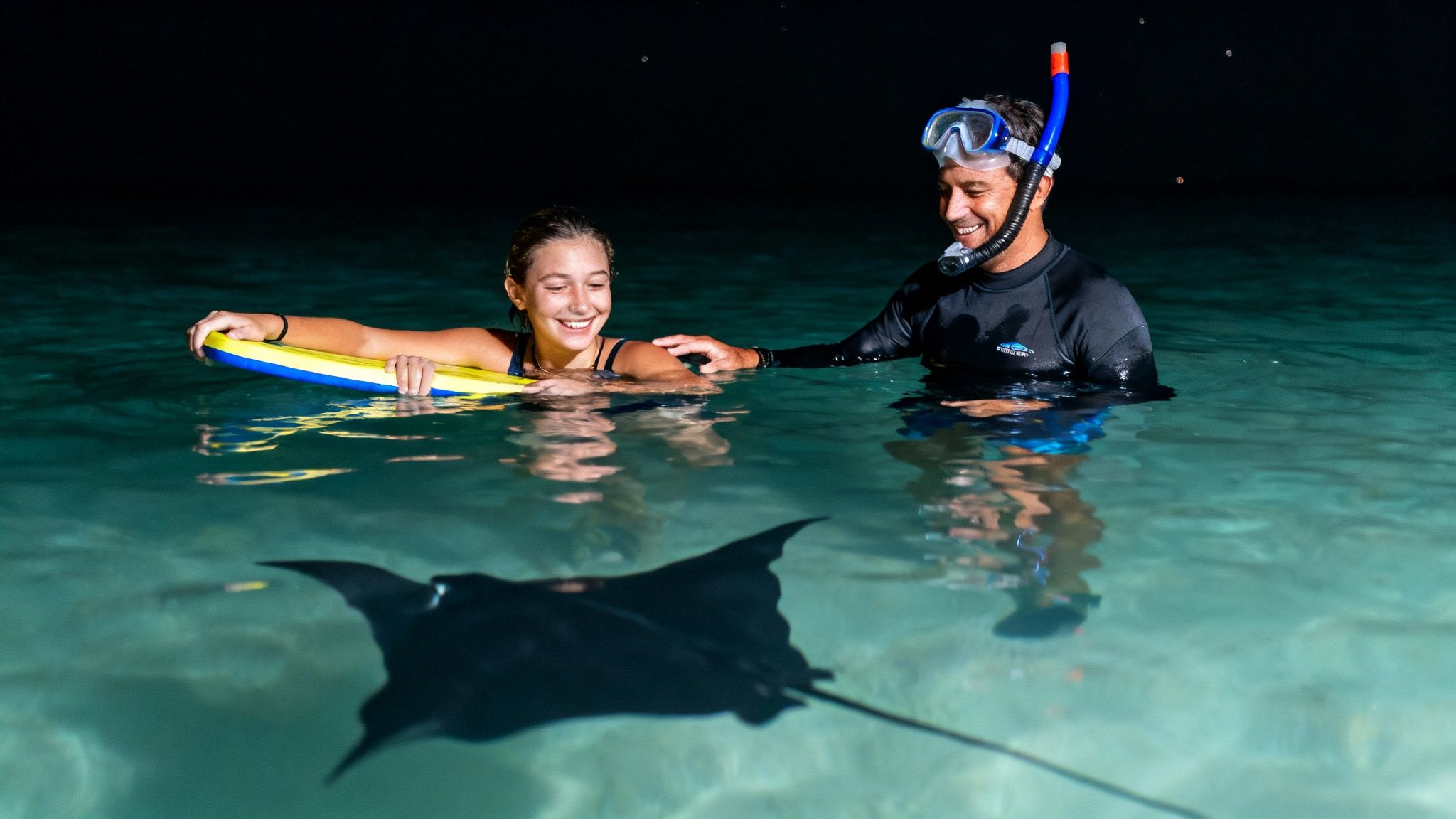 Two smiling people in clear water at night, observing a manta ray during a snorkel.