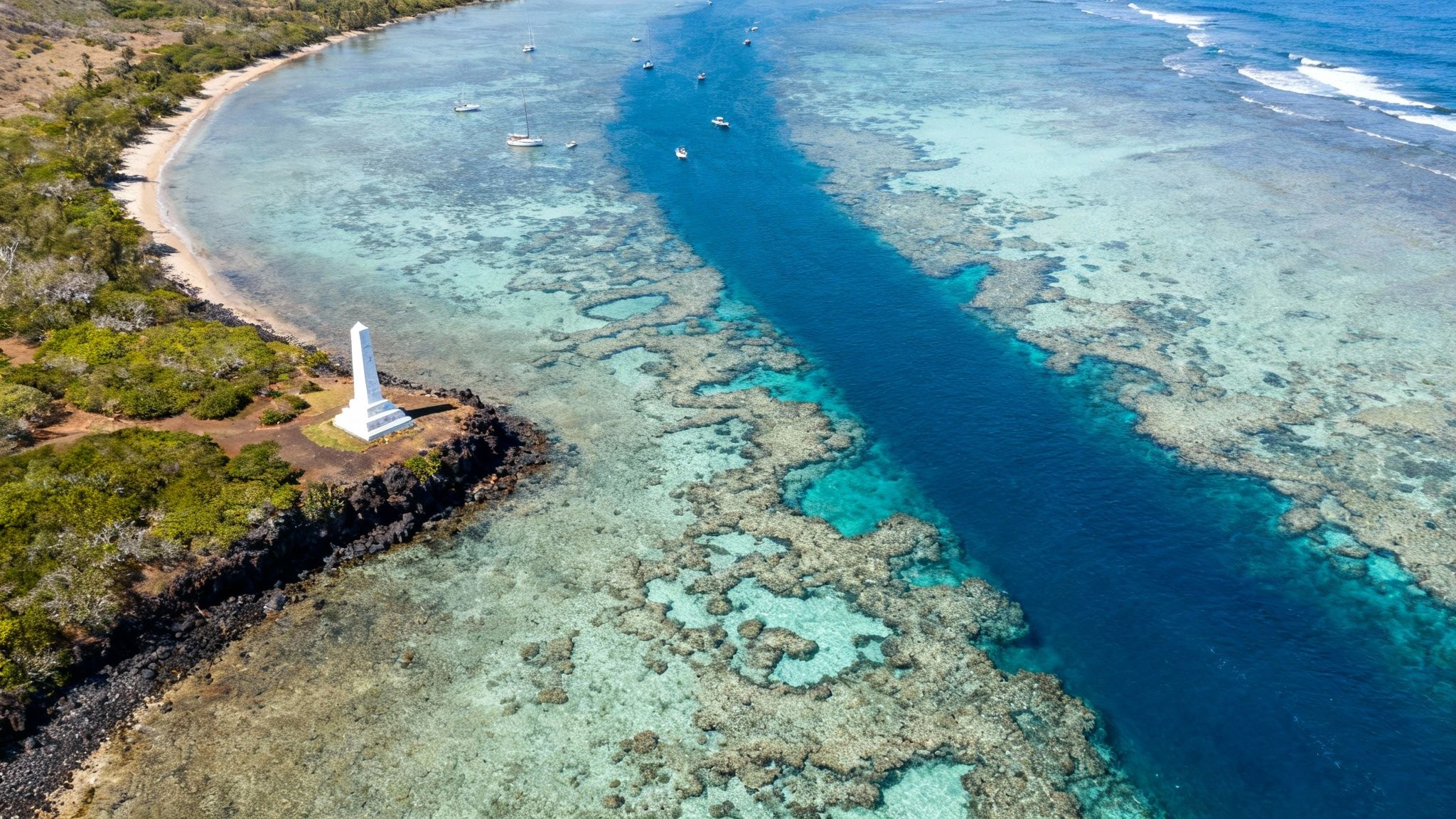 Aerial view of a white monument on a rocky point overlooking clear turquoise ocean with a coral reef and boats.