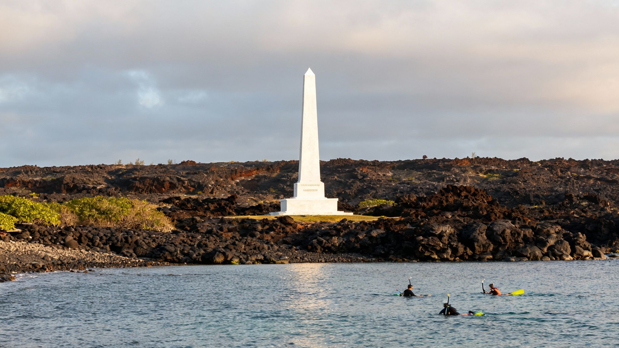 White Captain Cook monument obelisk stands among volcanic rocks with snorkelers in turquoise water
