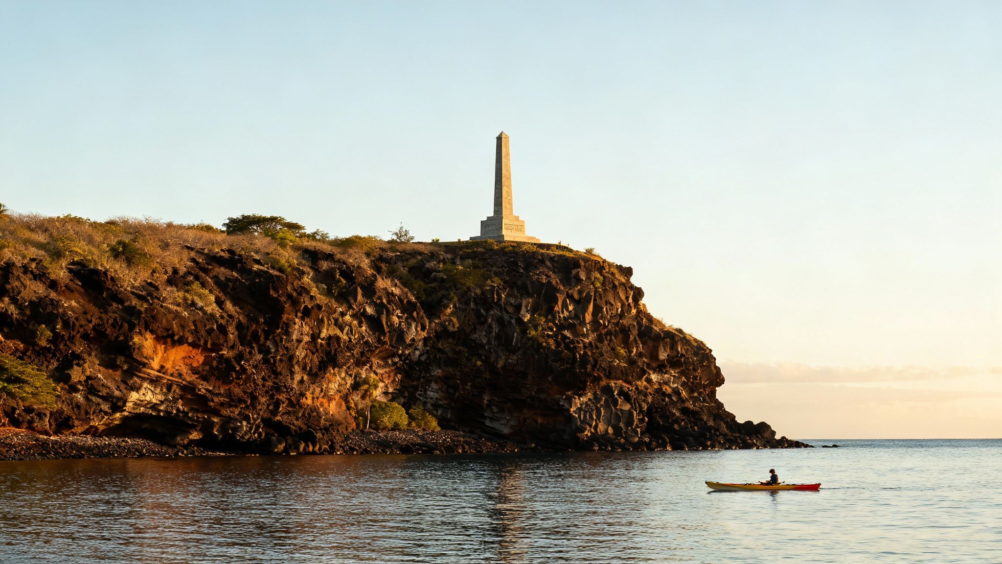 A historical monument, an obelisk, stands atop a rocky cliff overlooking a calm bay where a kayaker paddles.