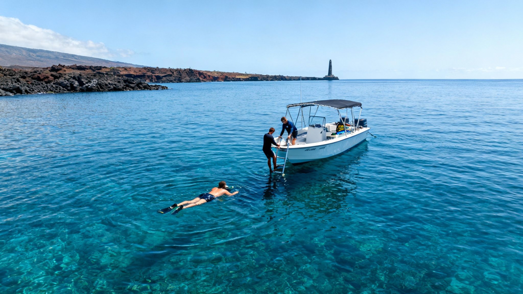 Snorkeler swims in clear blue water near a boat with two people on board, rocky coast, and a lighthouse.