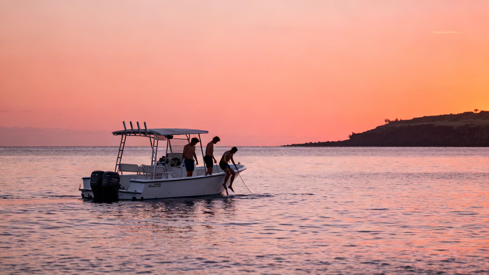 Three men on a white boat at sunset, one dipping feet into the calm, rosy ocean.