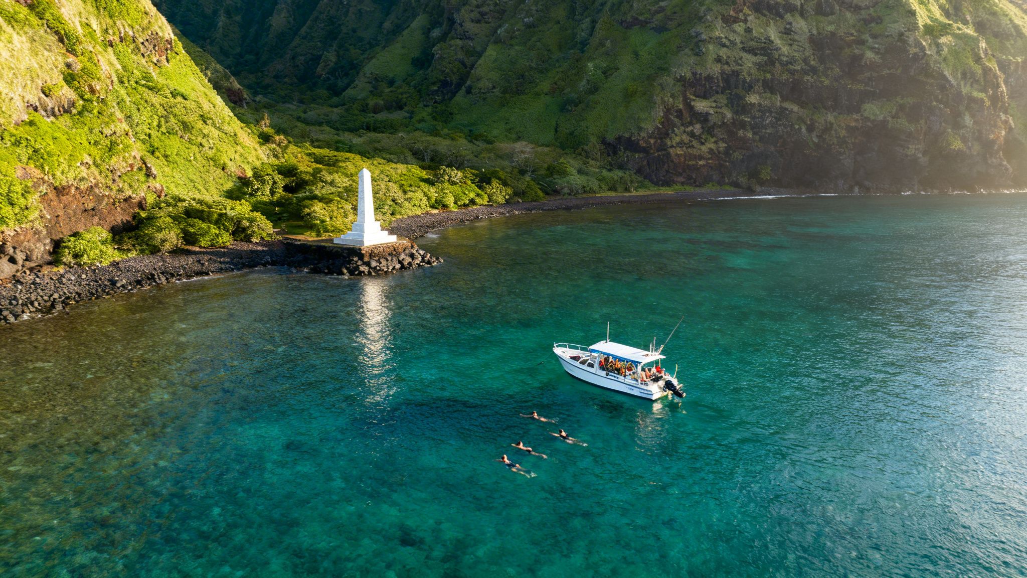 Aerial view of a boat and snorkelers in clear turquoise water near a white monument and green cliffs.