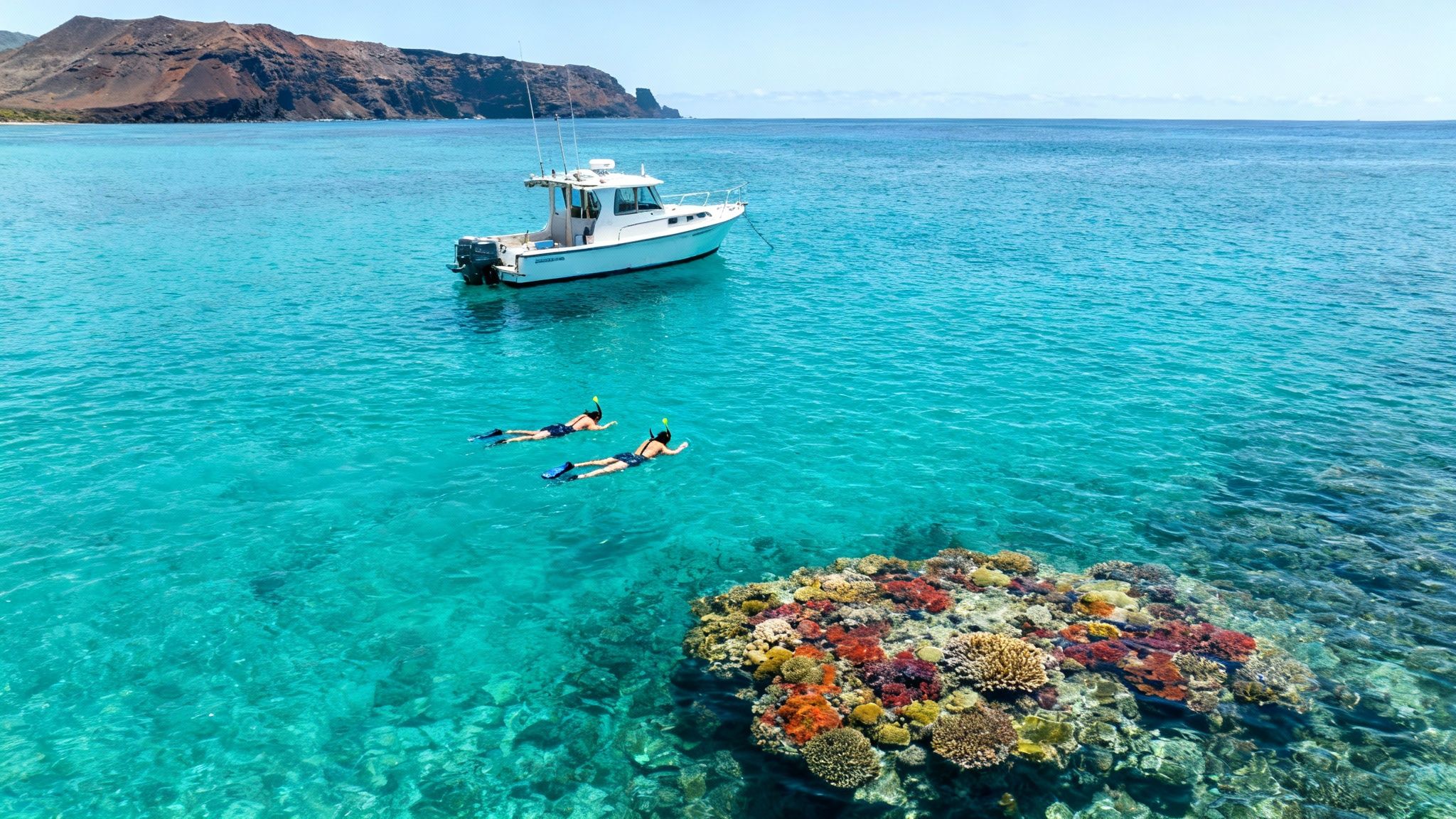 Two people snorkel near a vibrant coral reef in clear turquoise water, with a boat and island.
