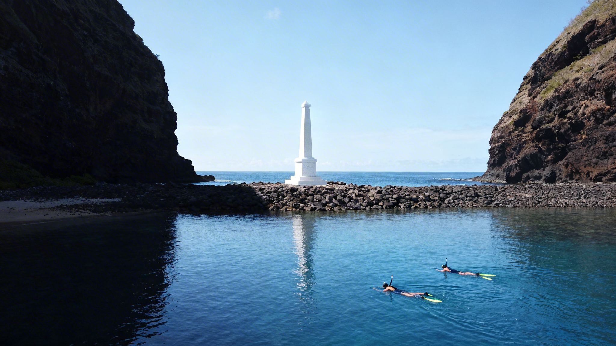 Two snorkelers enjoy clear blue waters near a white ocean monument framed by dark cliffs.