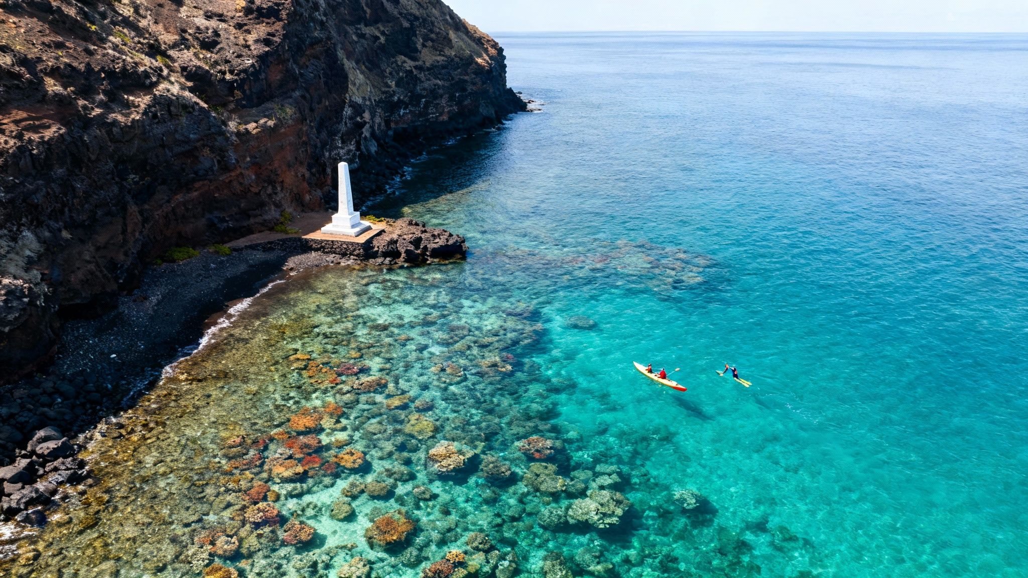 Aerial view of a white monument on a rocky beach, clear turquoise water, coral reefs, kayakers, and a snorkeler.