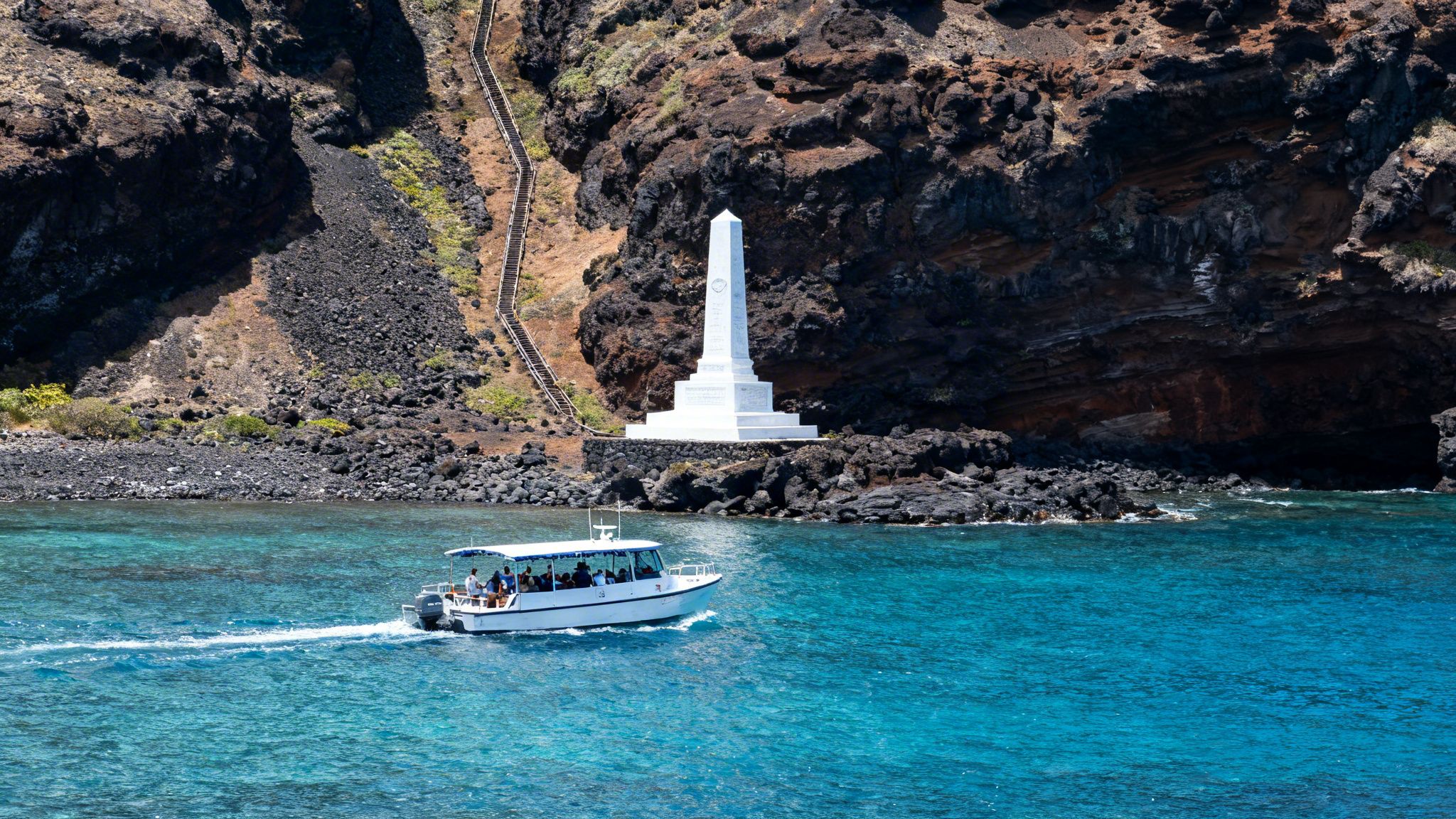 A white tour boat cruises past a rocky Hawaiian coastline with a white Captain Cook monument and stairs.