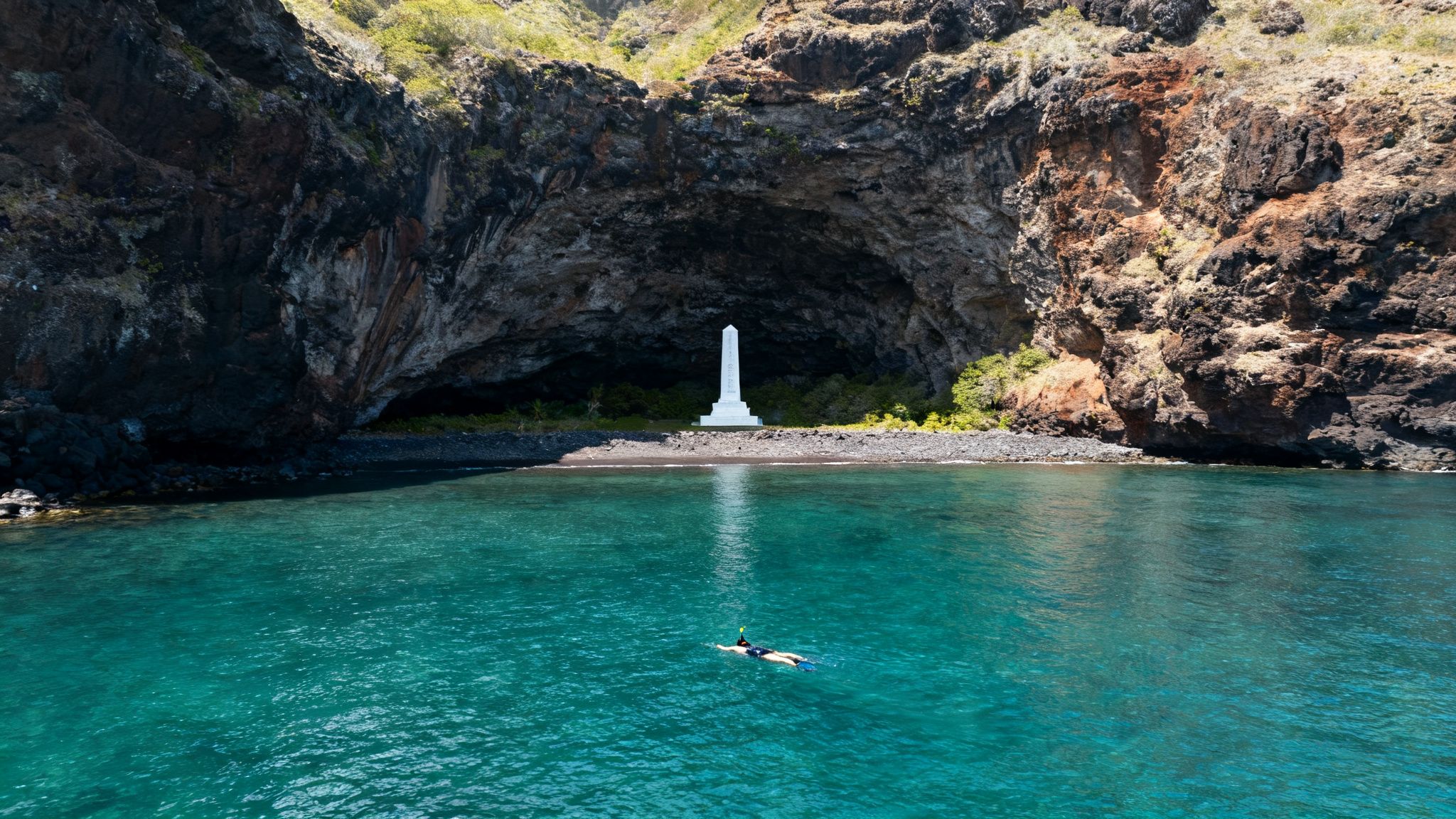 A person snorkeling in turquoise water towards a white monument on a beach inside a large sea cave.