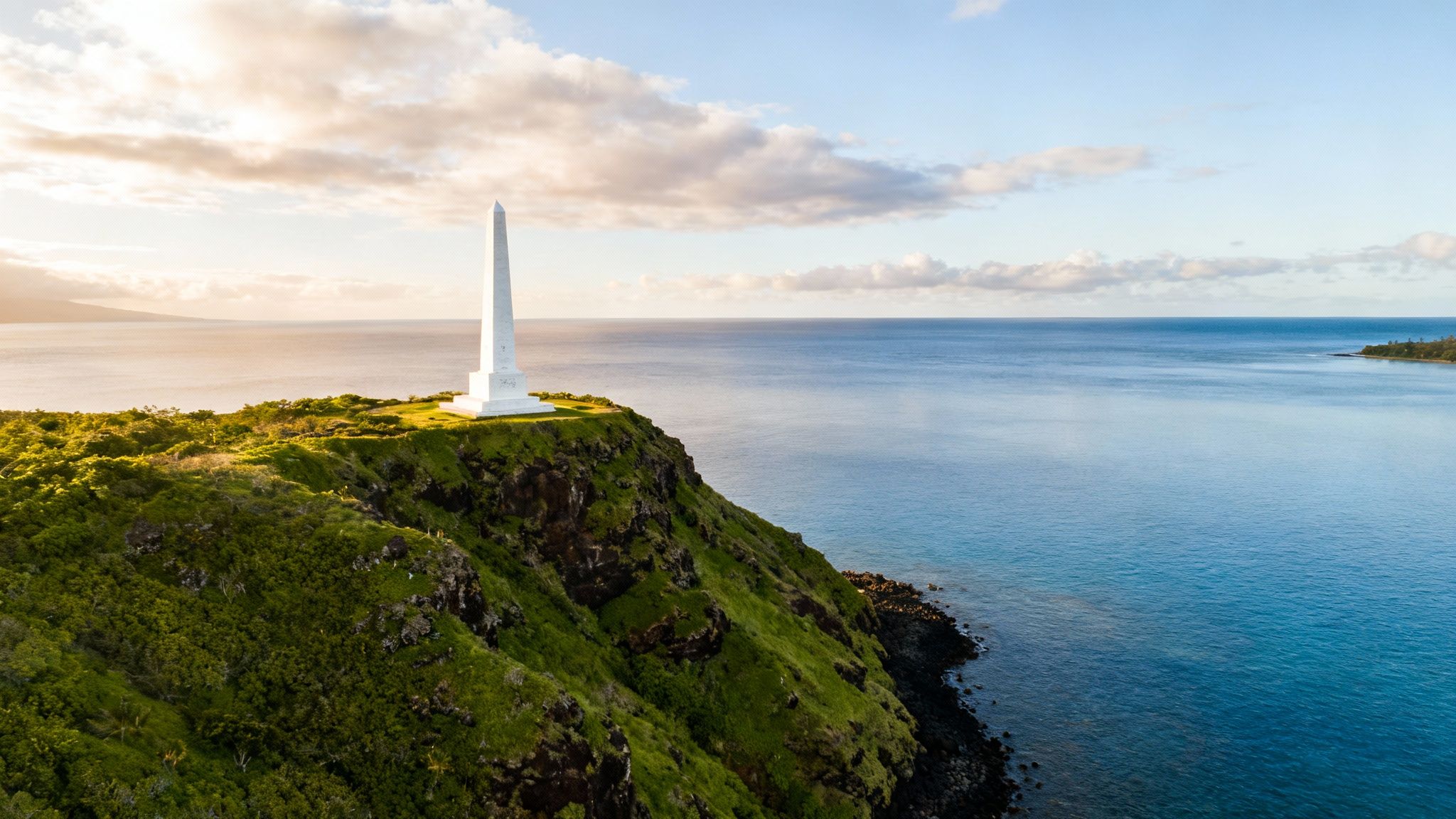 A white obelisk monument stands tall on a lush green cliff overlooking a calm blue ocean.