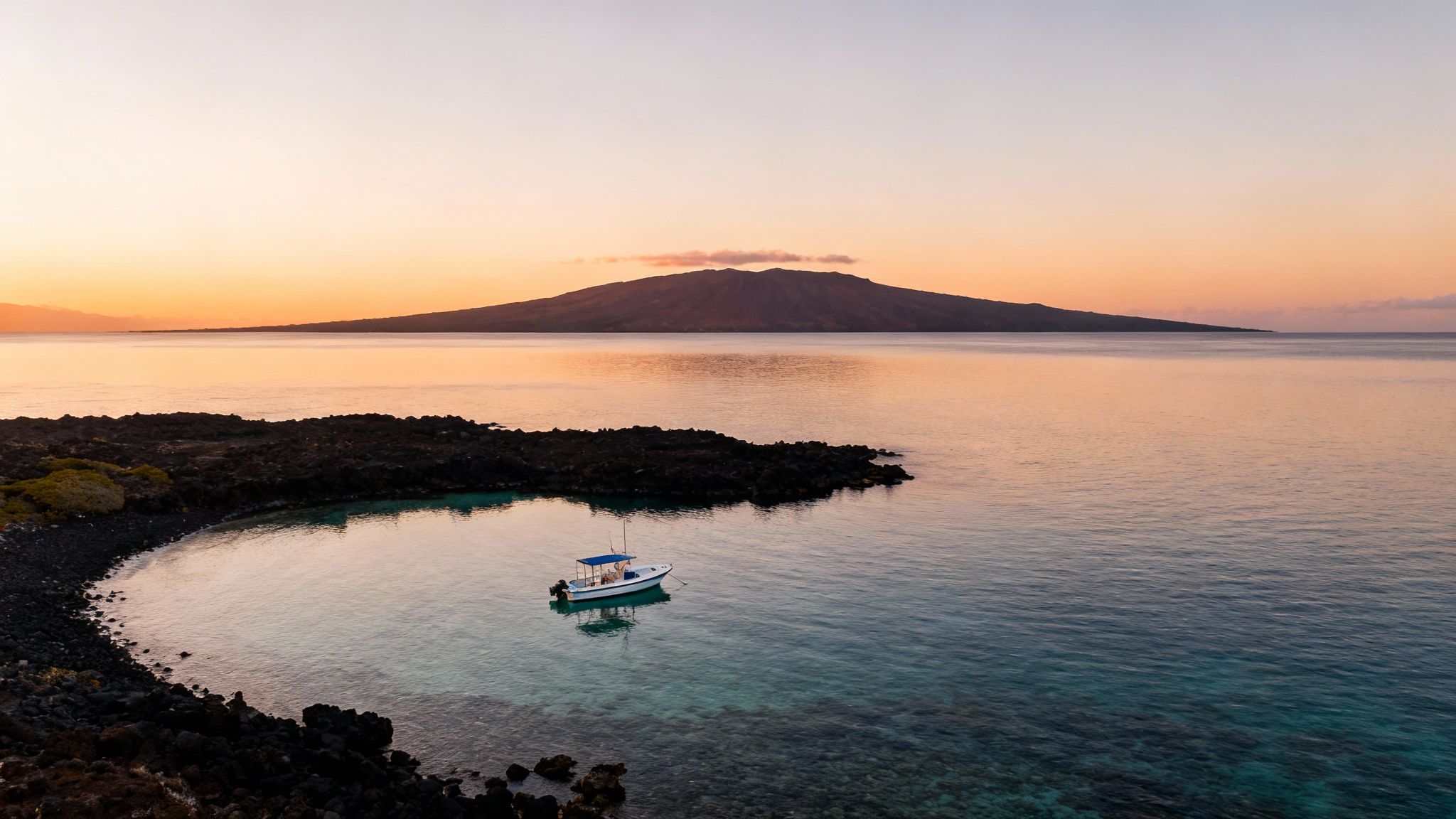 A small boat anchors in a clear, rocky cove at sunrise, with a large island in the distance.