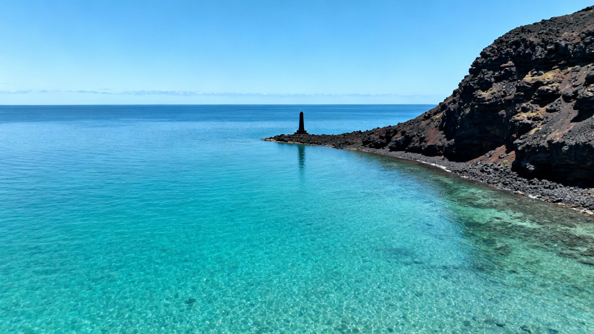 Aerial view of clear turquoise ocean water, a dark volcanic coast, and a black monument.