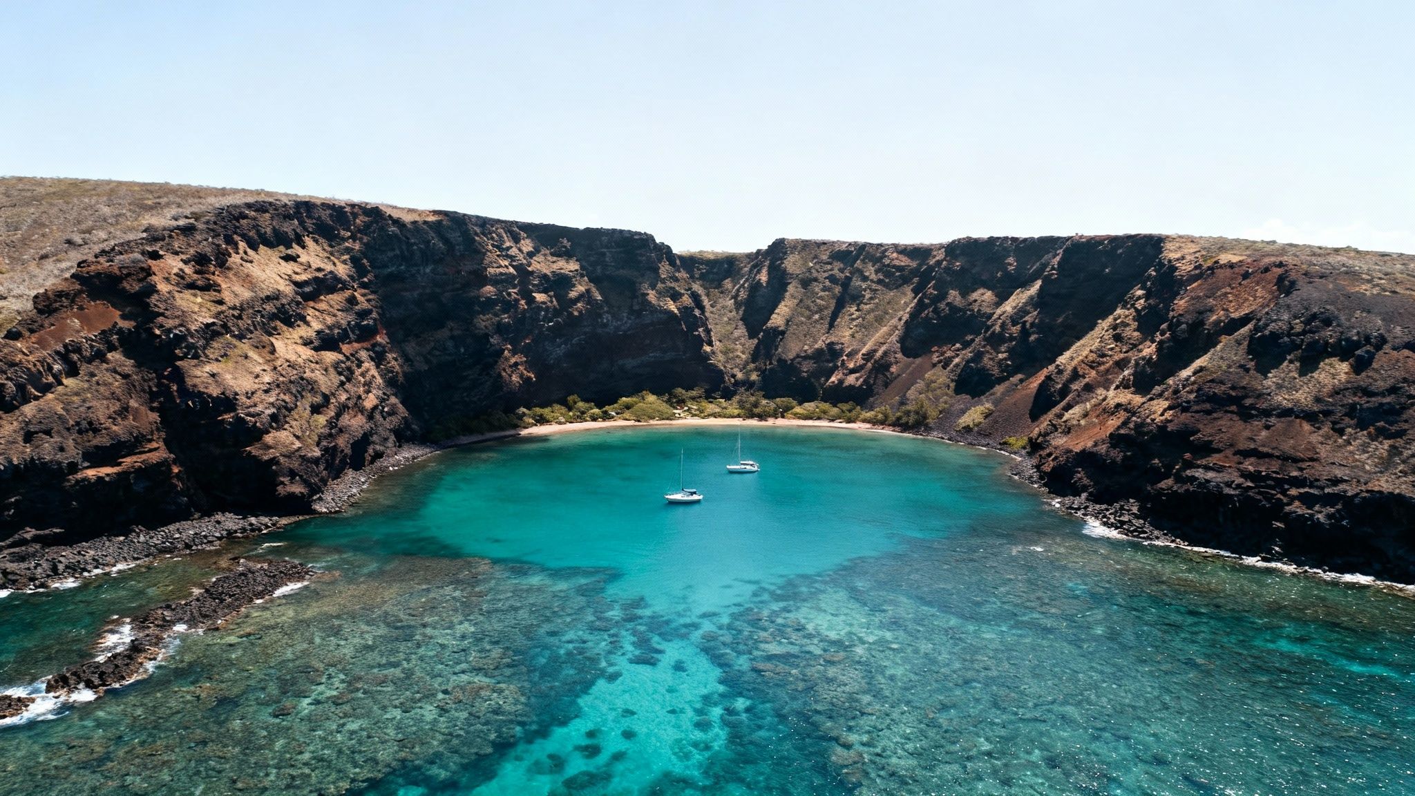 Aerial view of a serene bay with clear turquoise water, surrounded by dramatic volcanic cliffs, two sailboats anchored.