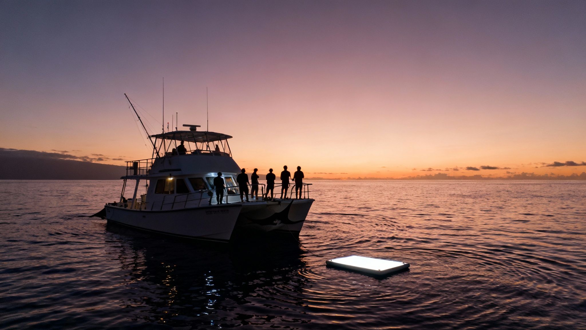 Silhouetted people on a catamaran at sunset, with a glowing light panel for night snorkeling.