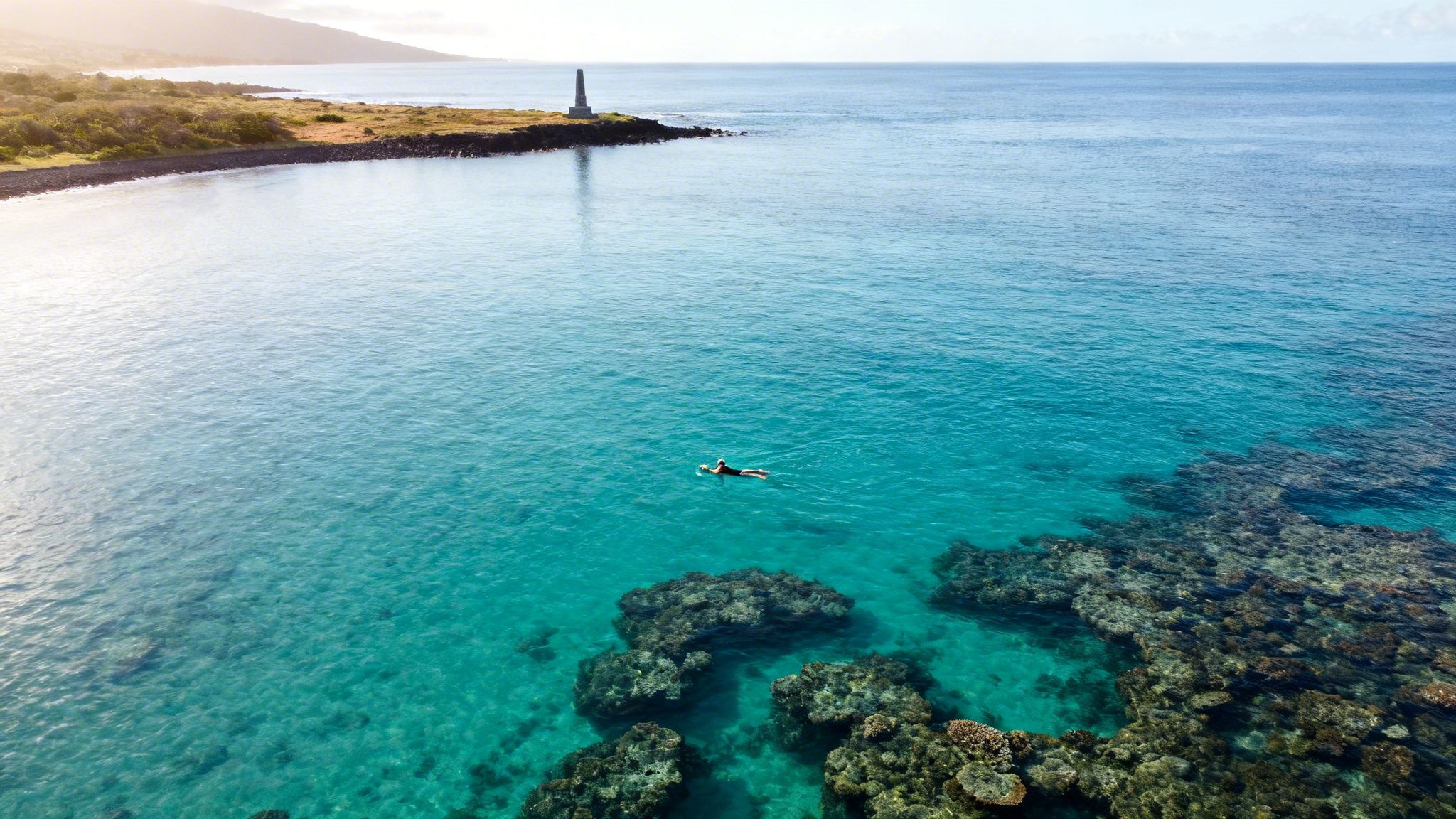Aerial view of a person paddleboarding in clear turquoise ocean water near a lighthouse and coral reefs.