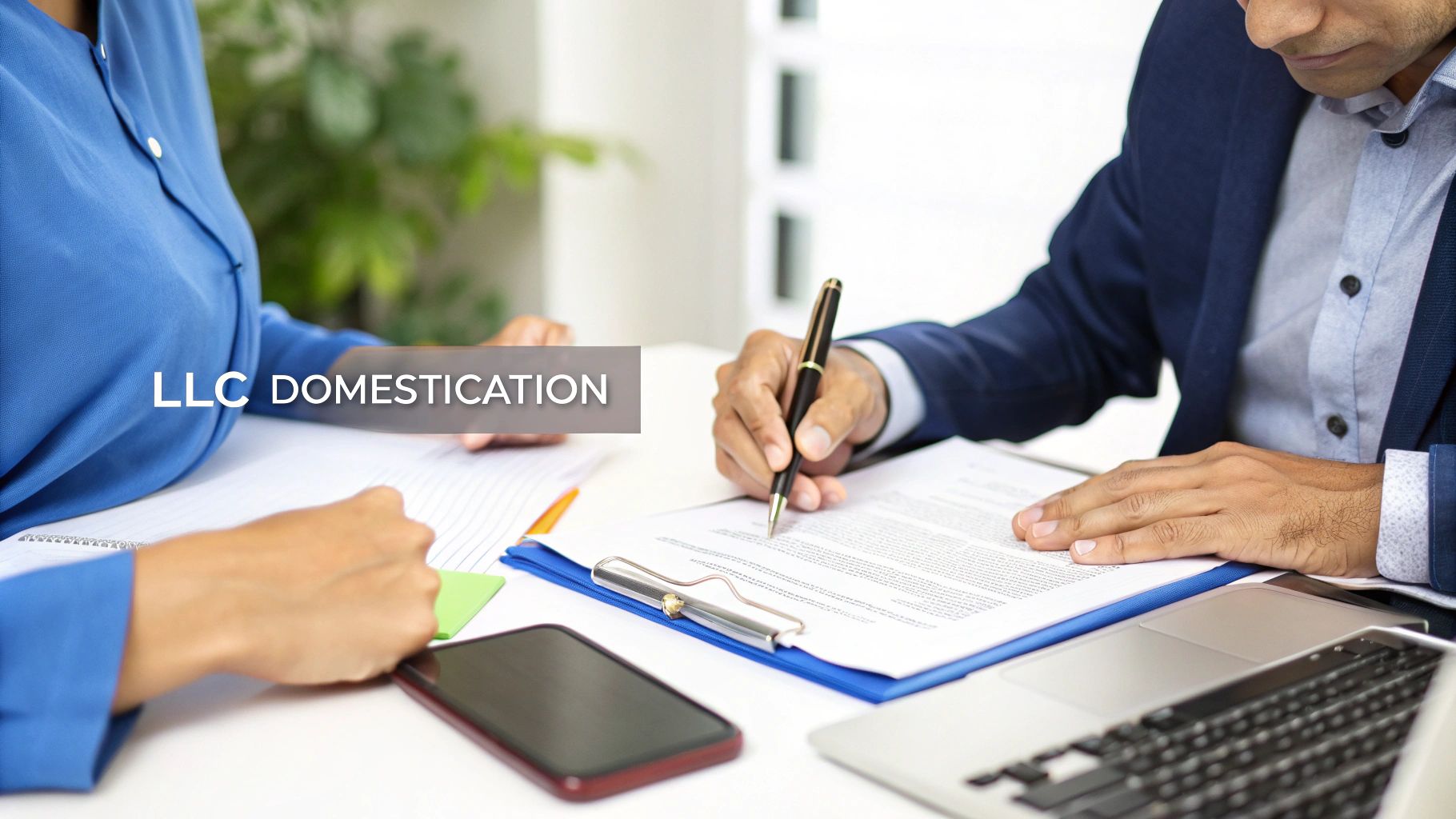 Two business professionals at a desk, one signing LLC domestication documents with a pen.