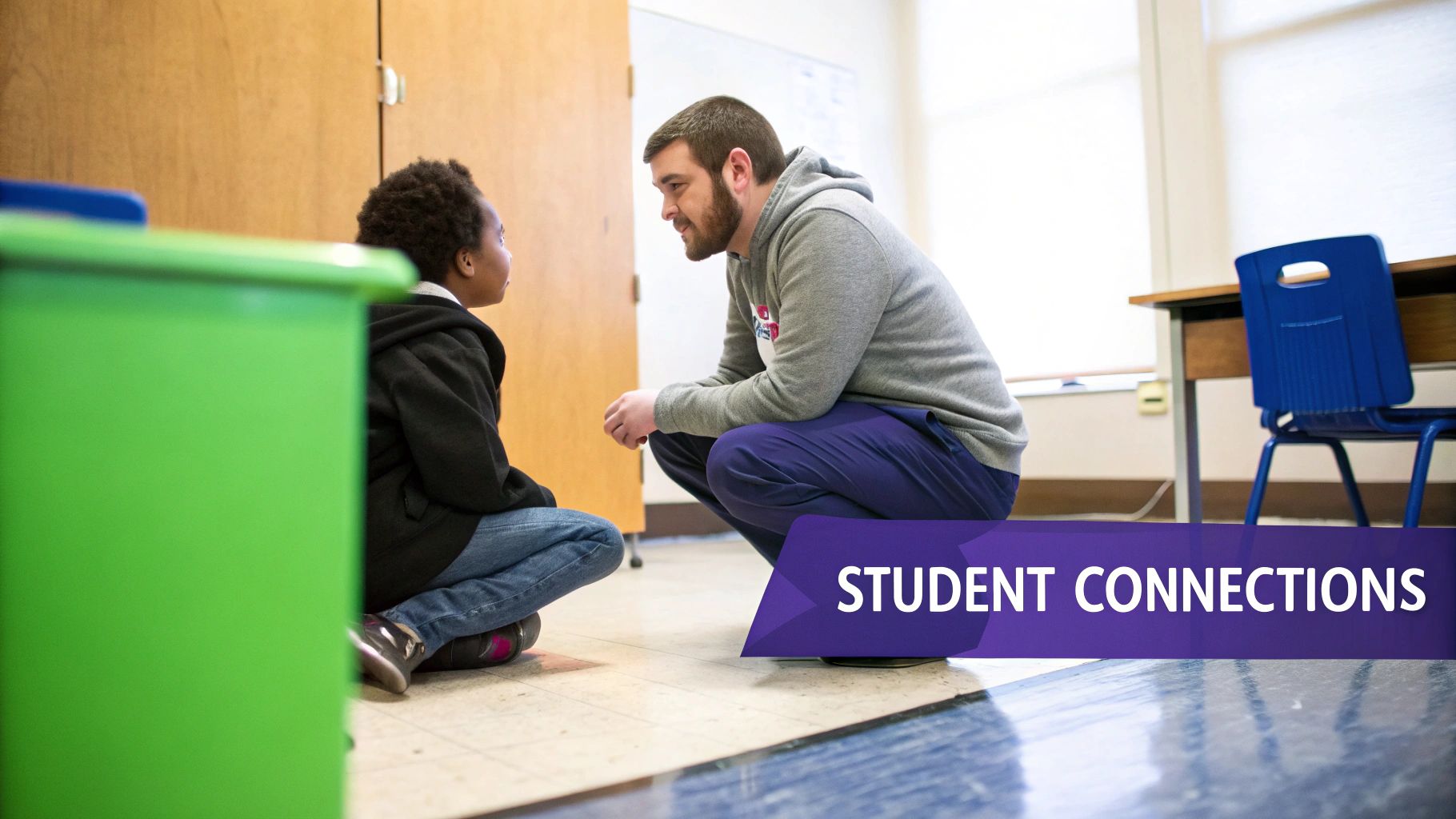 An adult male and a child crouch, facing each other and talking in a classroom.