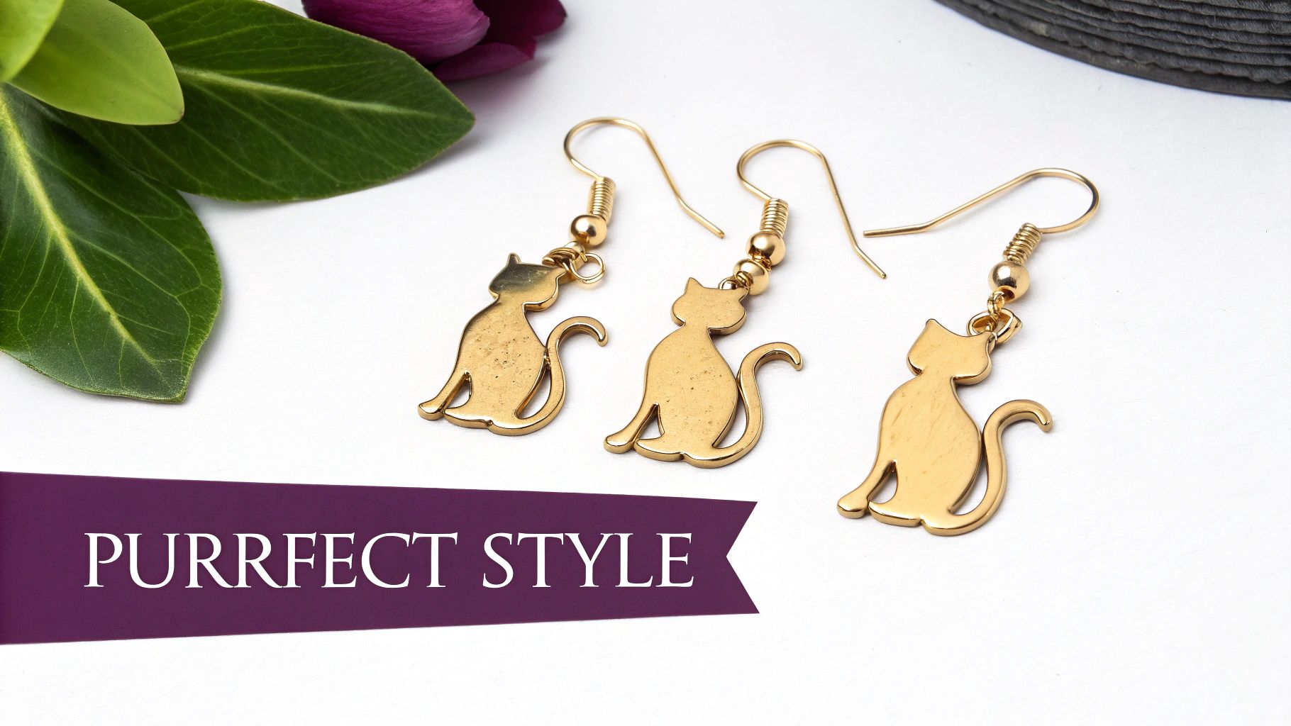 Three golden cat-shaped dangle earrings with hooks, displayed next to green leaves on a white background.