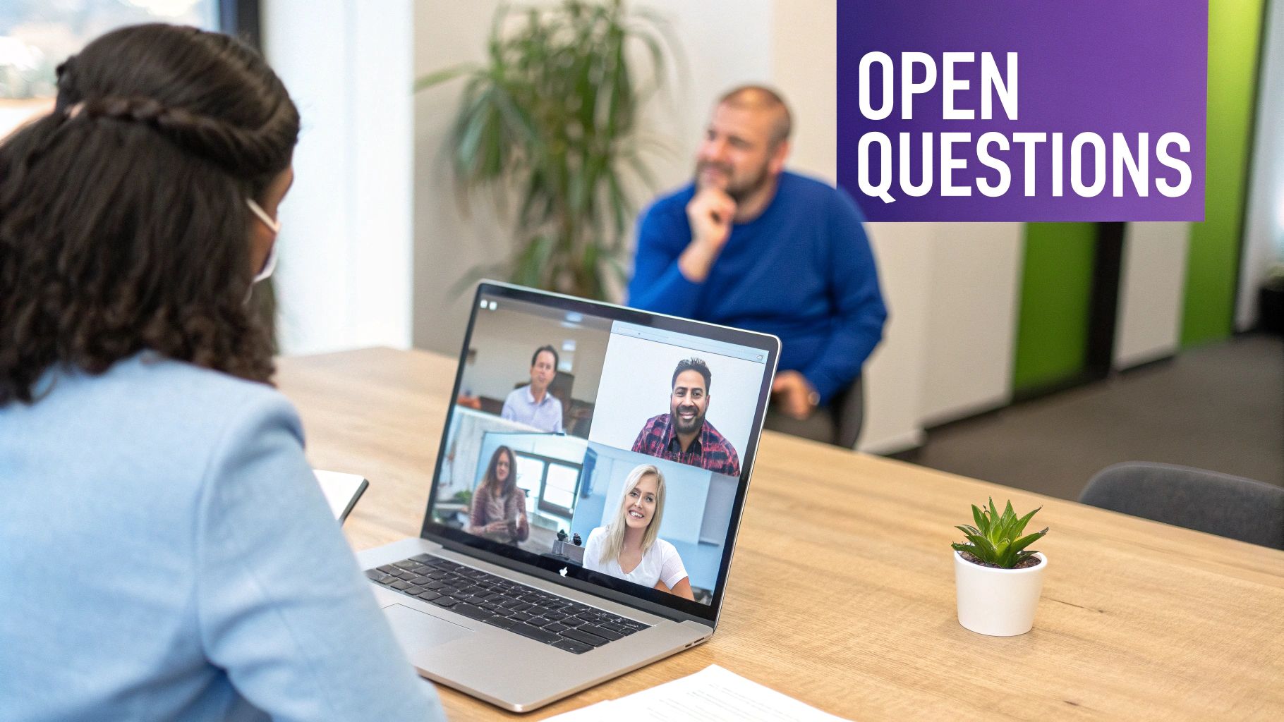 Person viewing a video conference on a laptop with four participants and a colleague nearby.